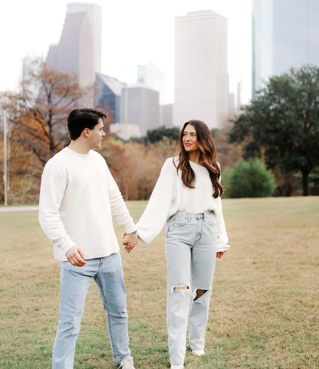 A young man and woman holding hands in a park with city skyline in the background