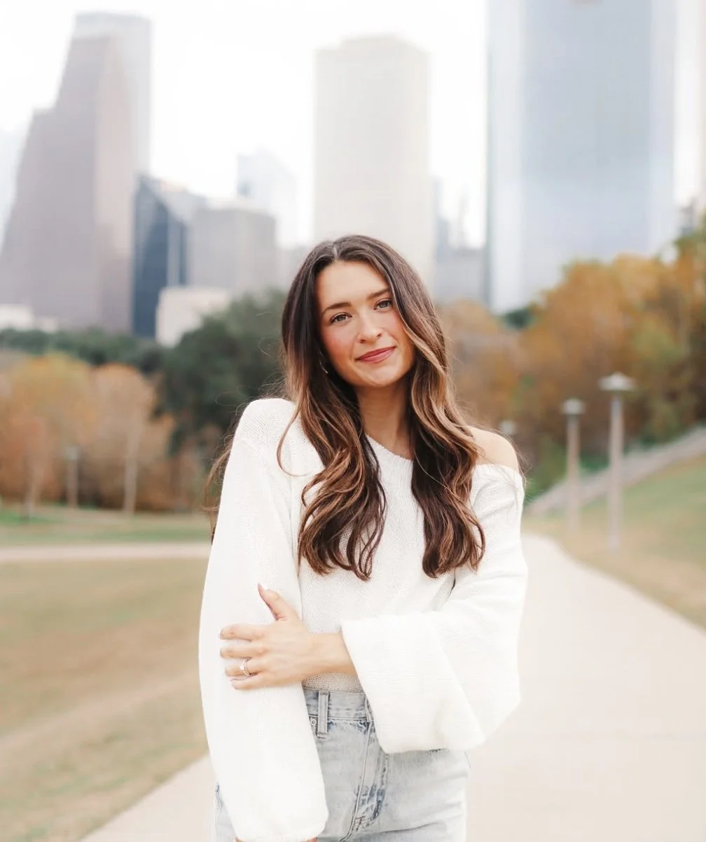 A young woman with long wavy brown hair wearing a white off-shoulder sweater and light jeans standing outdoors in an urban park with city skyscrapers in the background during autumn.