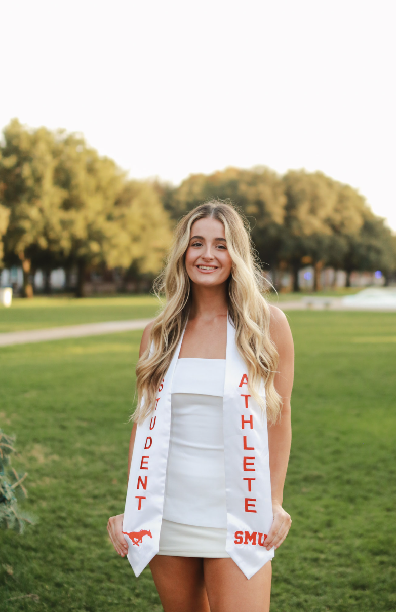 Young woman standing outdoors on grass, wearing a white dress and a sash that reads "Outstanding Athlete" with a Mustang logo and the abbreviation "SMU".