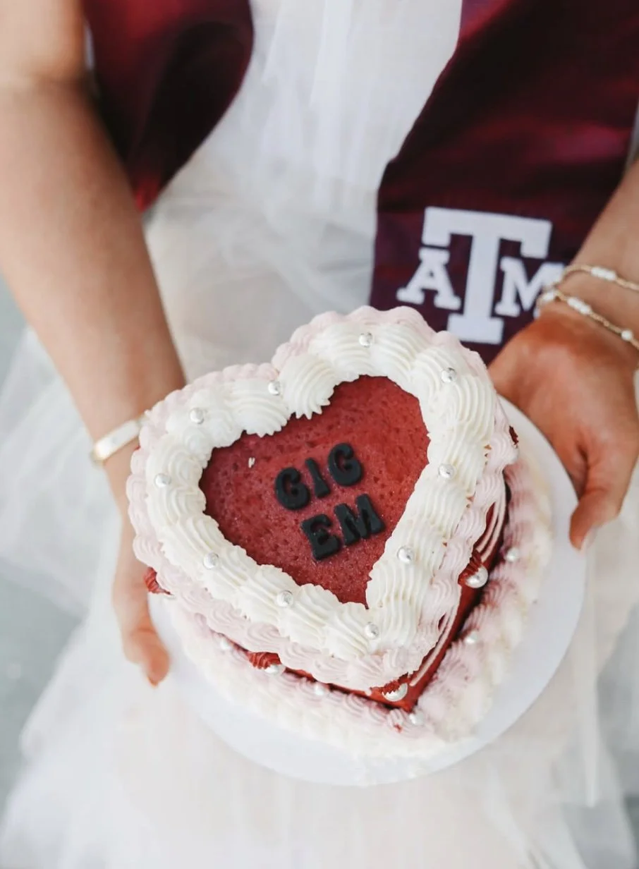 Close-up of a heart-shaped red velvet cake with white whipped cream frosting and silver decorations, with the words 'GIG EM' written on top in black letters, held by a person wearing a ring and bracelets, with a person in a Texas A&M University maroon and white shirt nearby.
