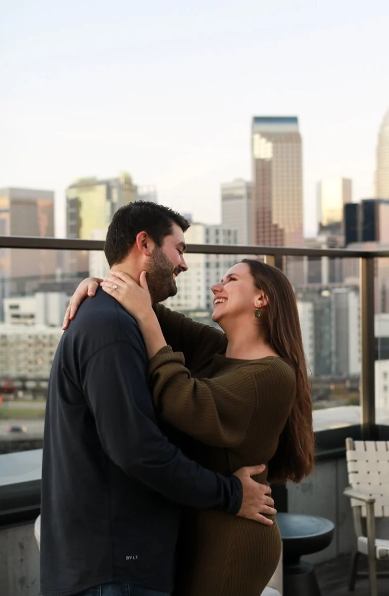 A couple sharing a joyful moment on a balcony with a city skyline in the background, embracing and smiling at each other.