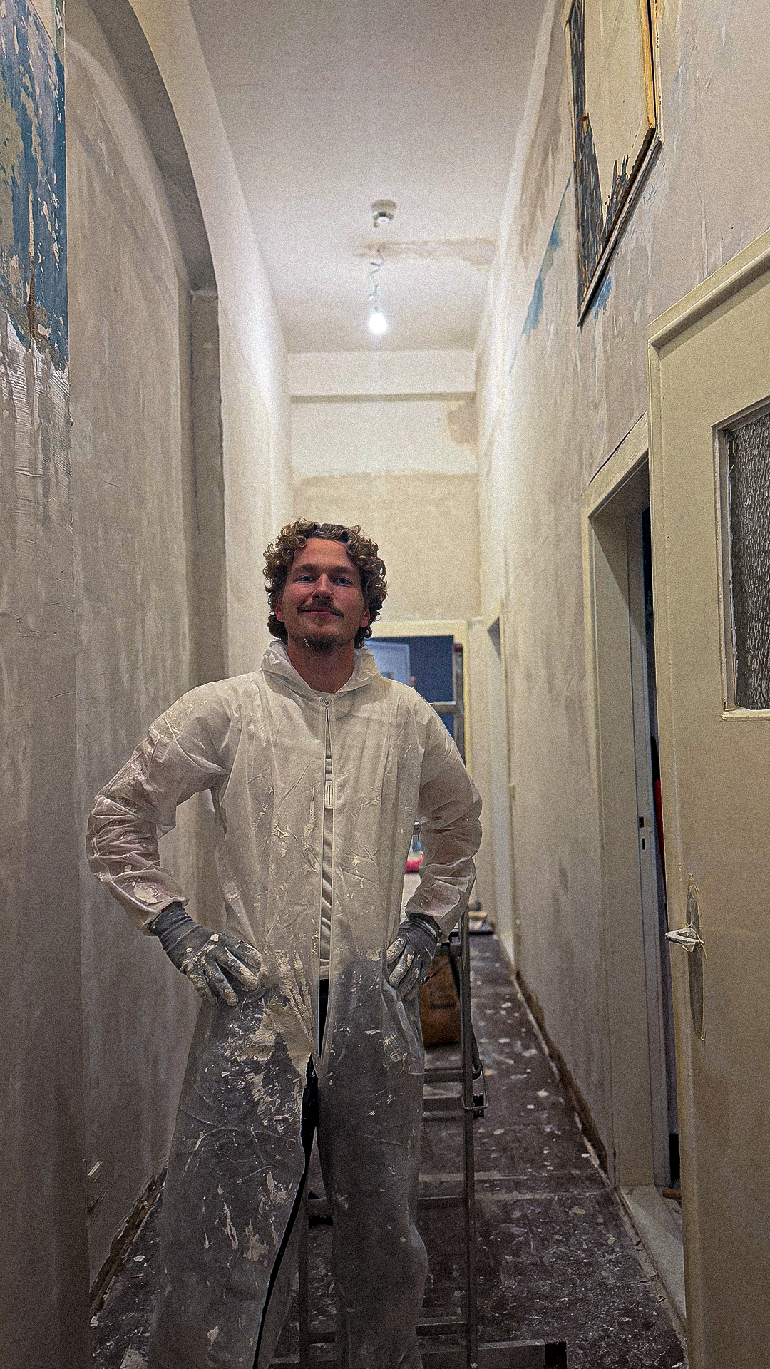 Ein Mann in weißen Maleranzug, Handschuhen und mit Spritzern von Farbe steht in einem renovierten Flur mit unvollständigen Wänden.