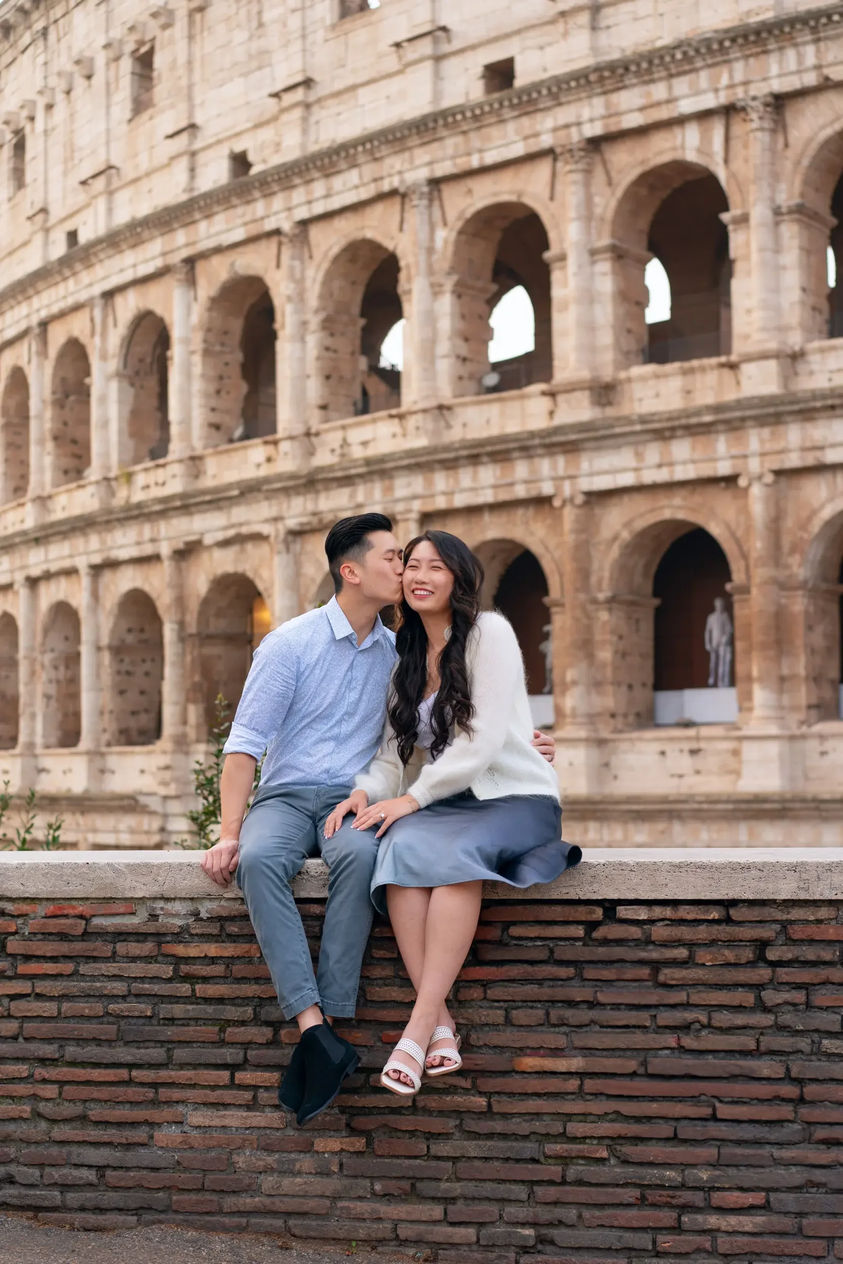 A couple sitting on a brick wall in front of the Colosseum in Rome, Italy. The man kisses the woman on the cheek as she smiles happily.