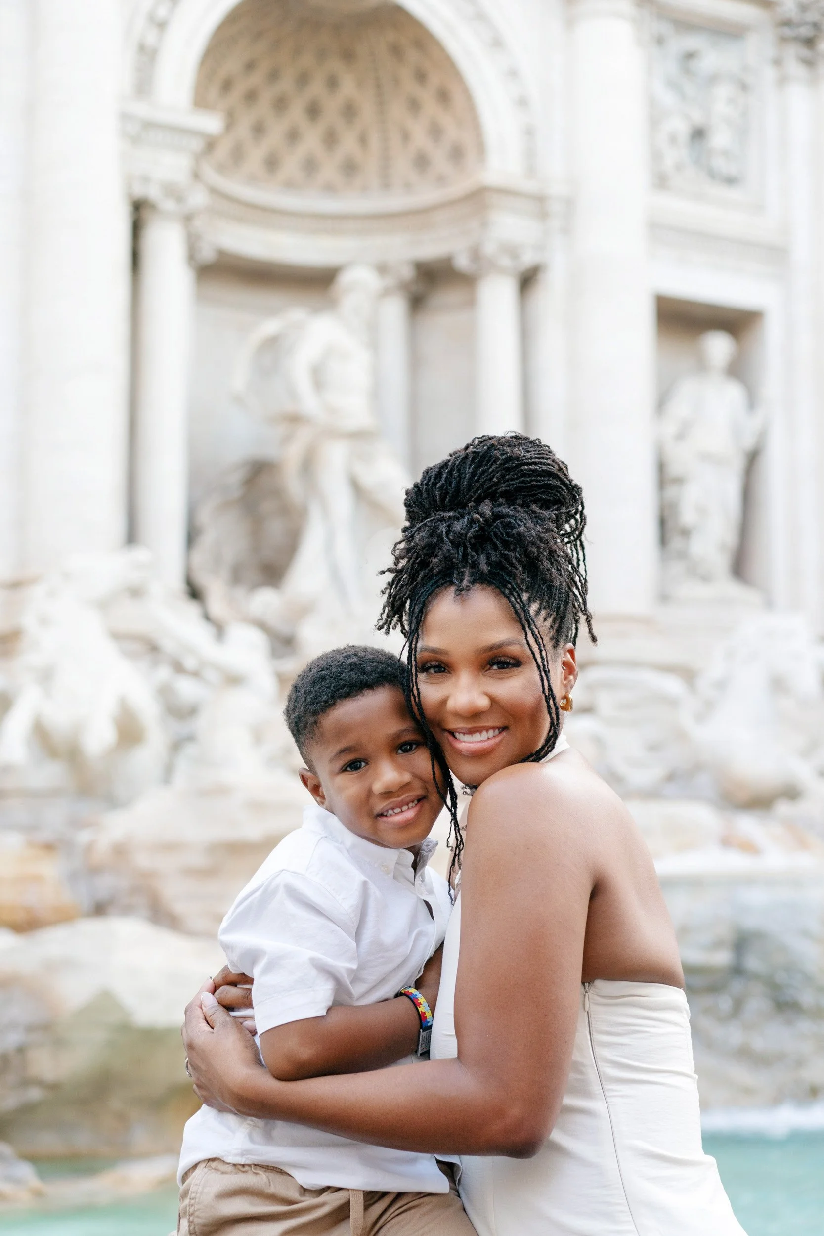 A woman and young boy smiling and hugging near a fountain with sculptures in the background.