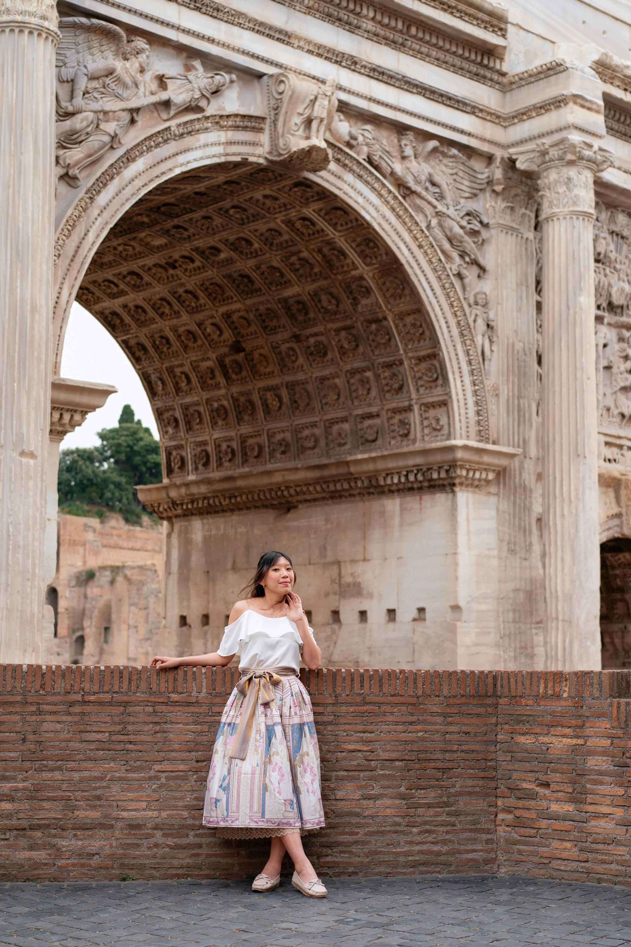 A woman in a white off-shoulder top and a patterned skirt stands against a brick wall in front of an ancient stone archway with intricate carvings.