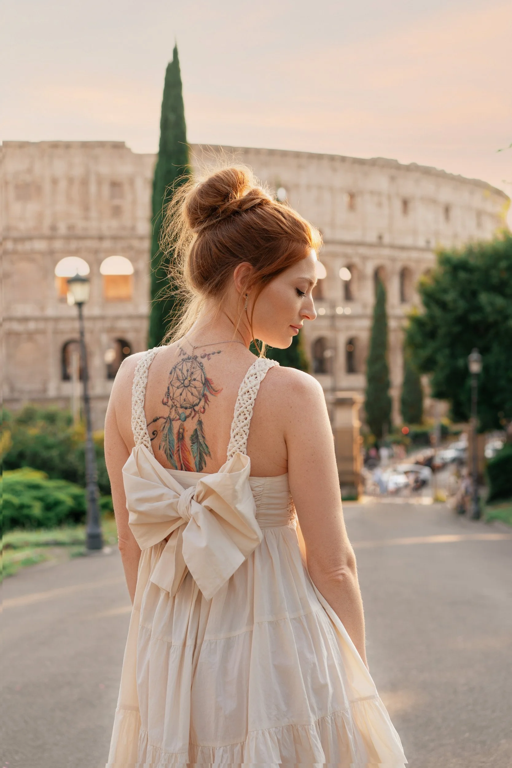 A woman with red hair in a bun, wearing a white dress with lace straps and a large bow on the back, standing outdoors near the Colosseum in Rome, Italy, during sunset.