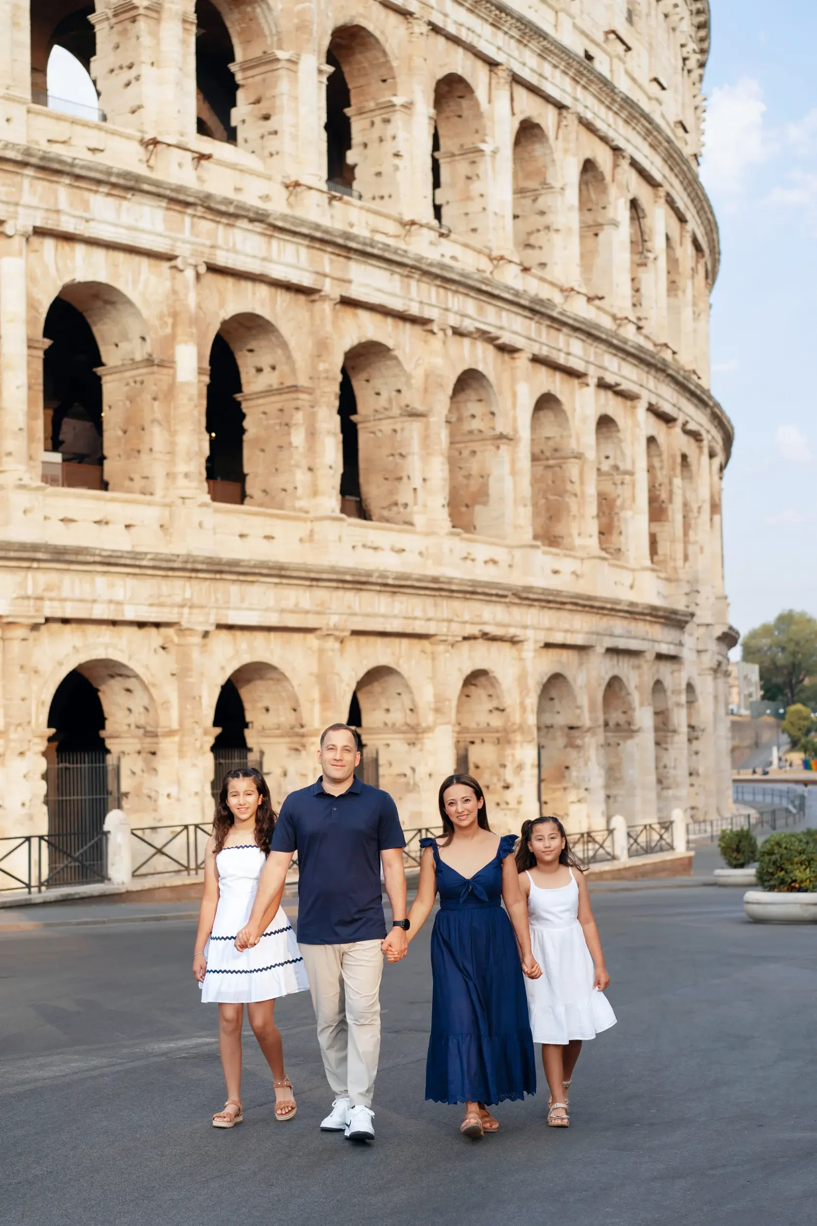 A family of four holding hands and walking in front of the Colosseum in Rome, Italy. The family includes two girls, a man, and a woman, all dressed in summer clothing.