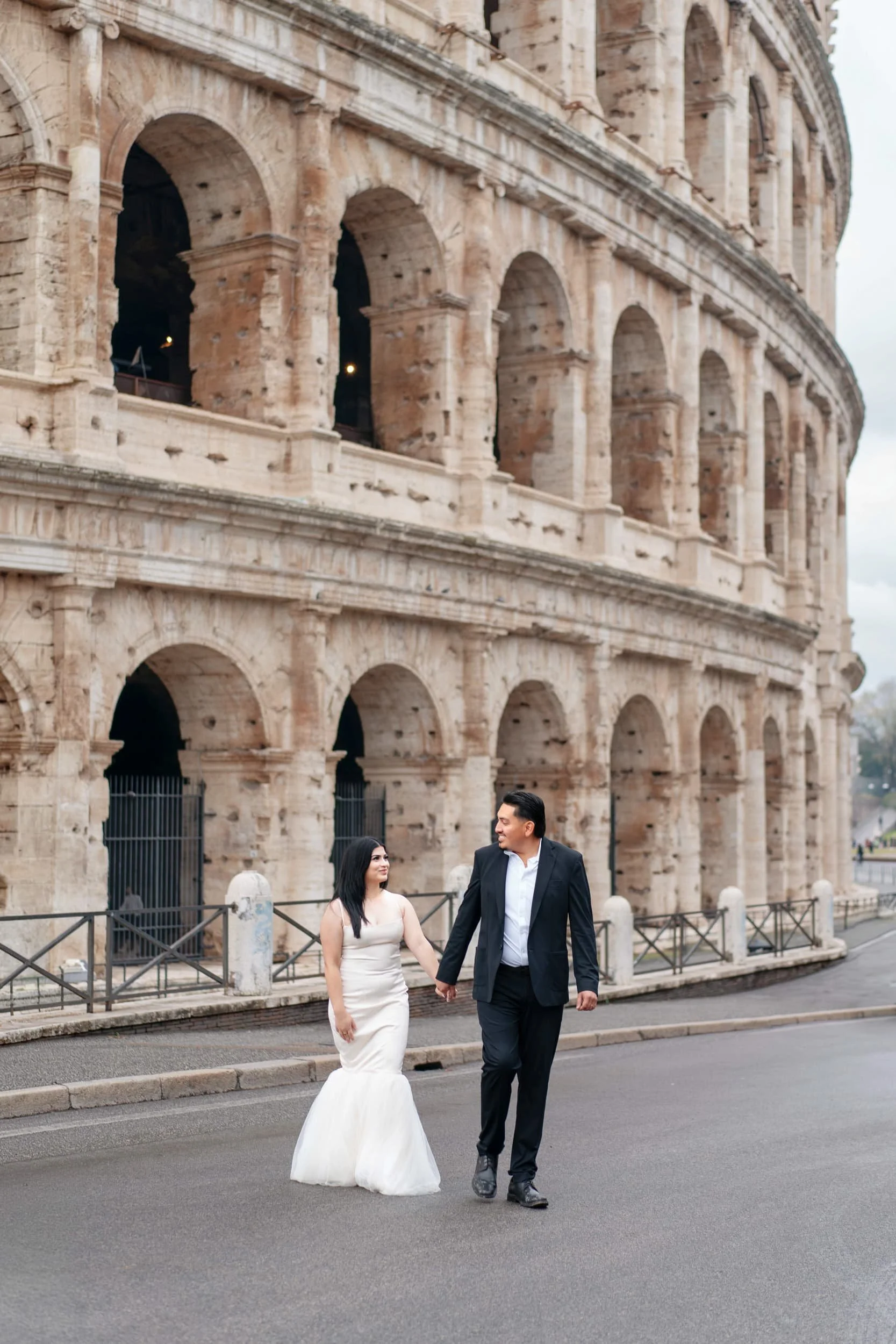 A couple in wedding attire holding hands and walking in front of the Colosseum in Rome, Italy.