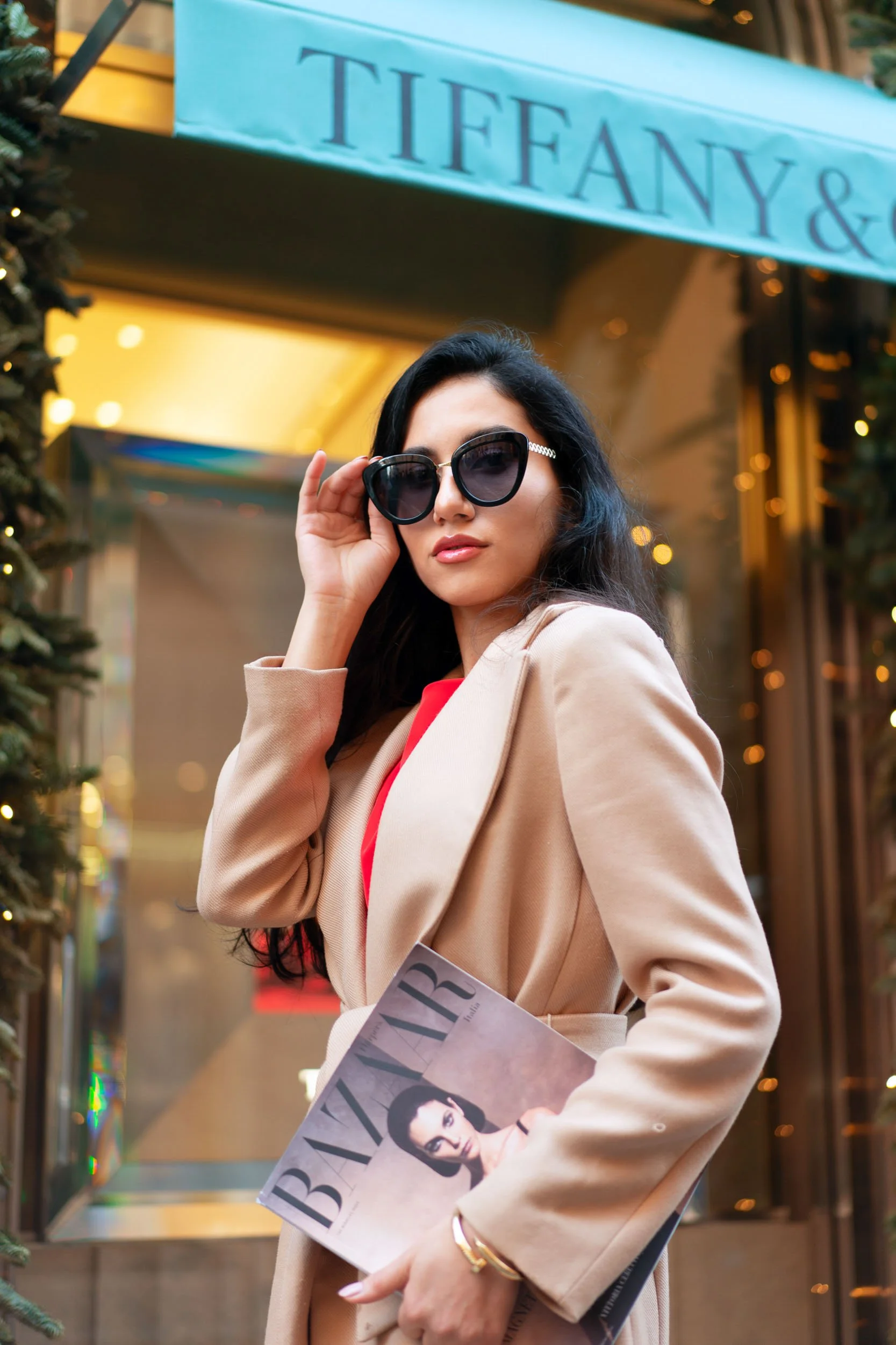 A stylish woman wearing large black sunglasses, a tan coat, and a red top standing outside a Tiffany & Co. store, holding a Harper's Bazaar magazine.