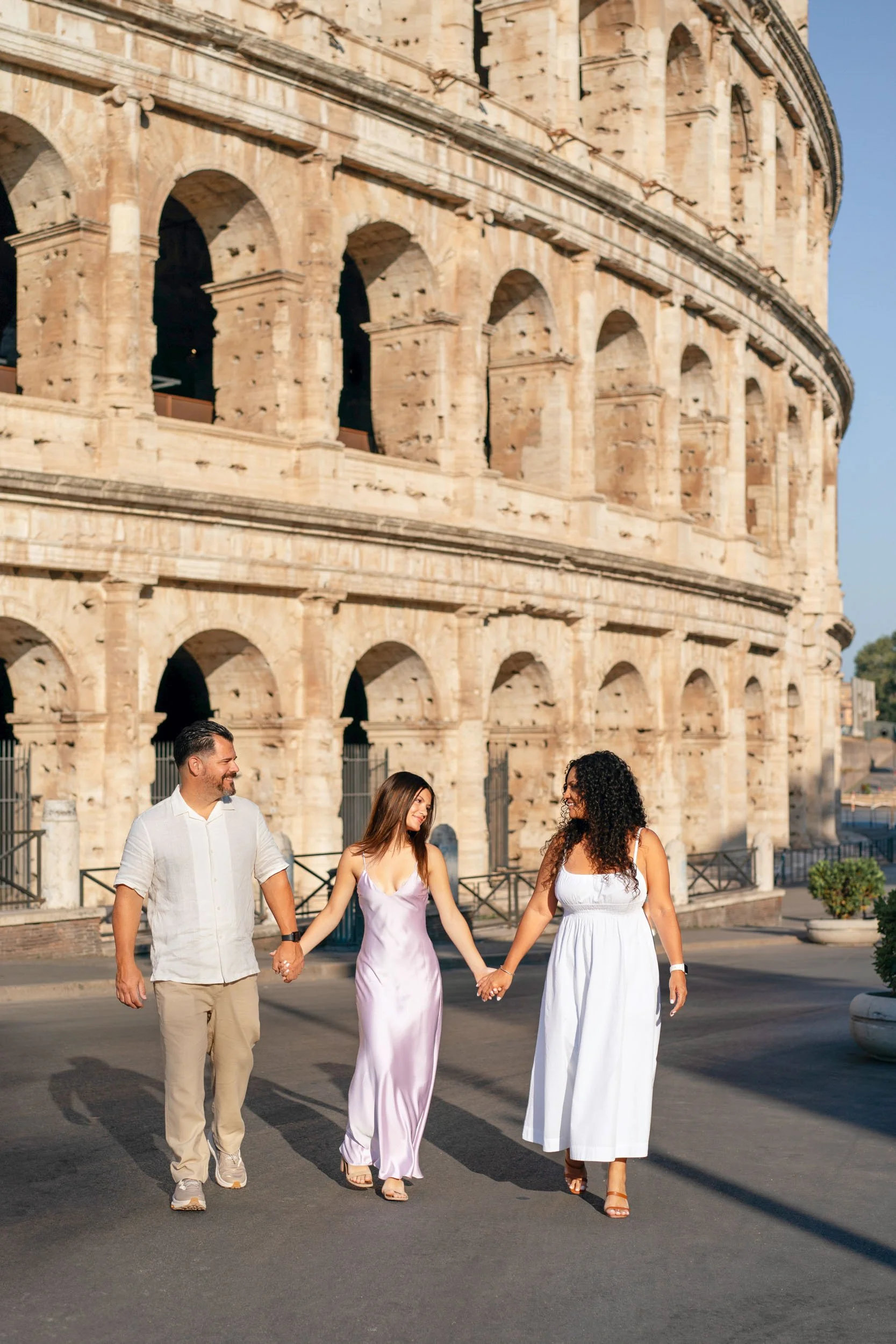 A group of three friends walking hand-in-hand in front of the Colosseum in Rome, during daytime, smiling and enjoying each other's company.