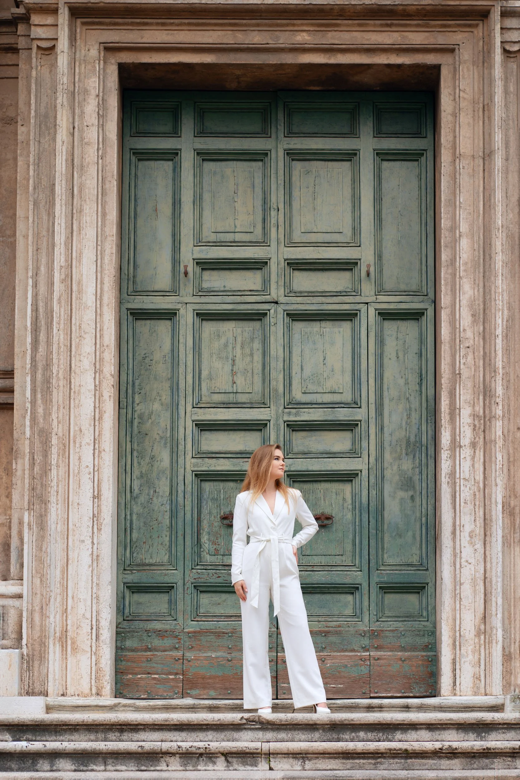 A woman in a white jumpsuit standing in front of a large green wooden door with a stone frame.