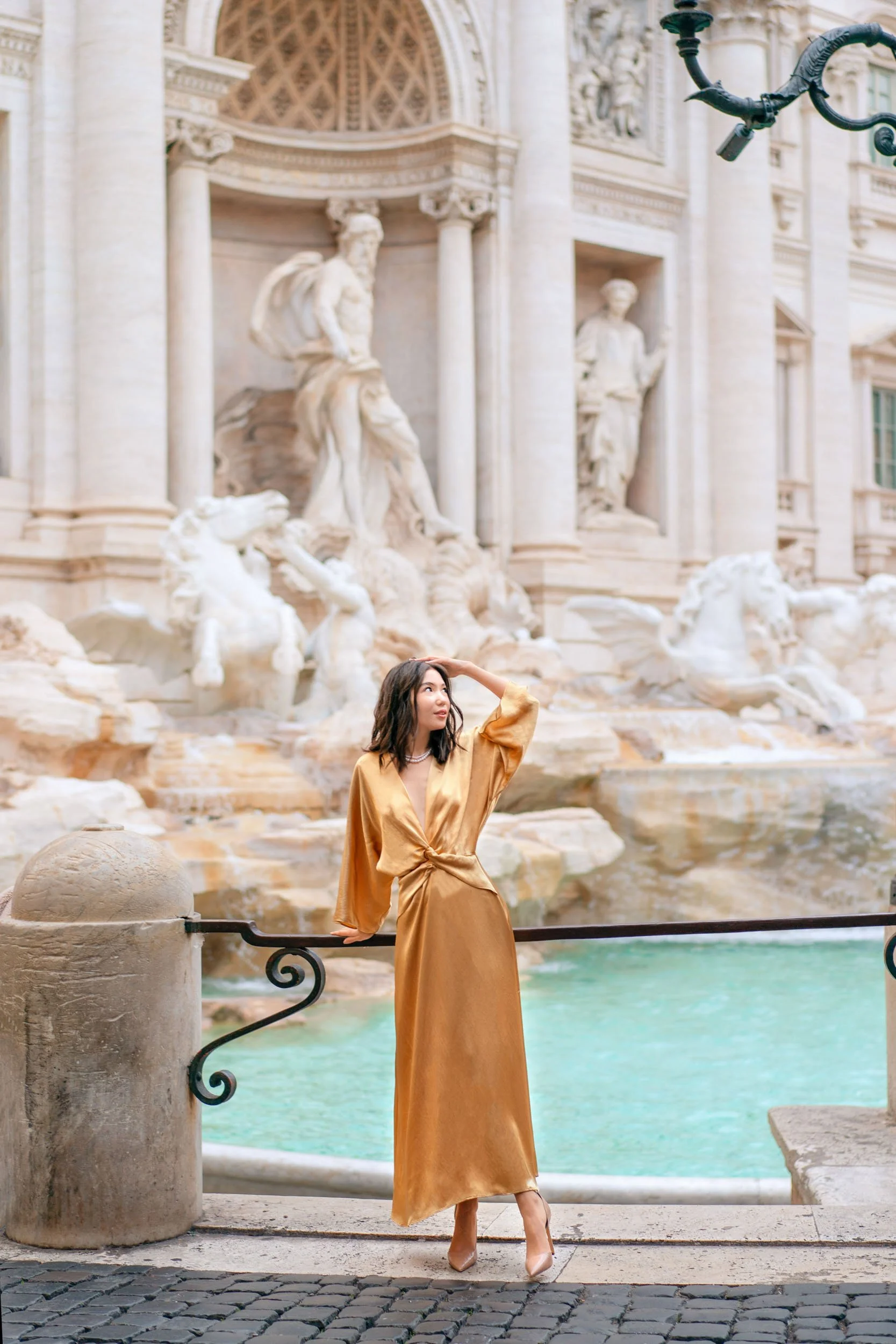 A woman in a gold satin dress standing by a fountain with sculptures in the background, looking to her left and touching her head.