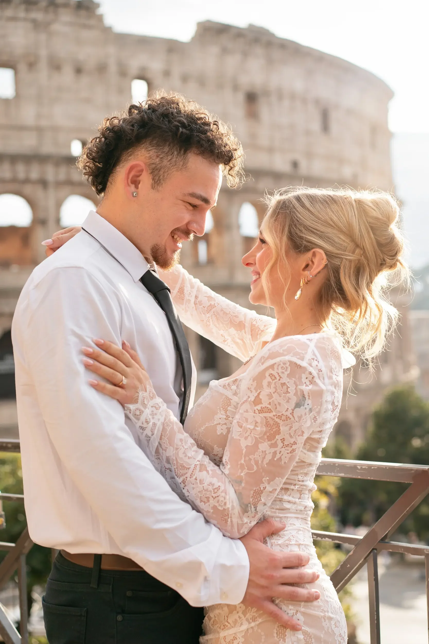 A couple in wedding attire smiling and holding each other in front of the Colosseum in Rome, Italy.