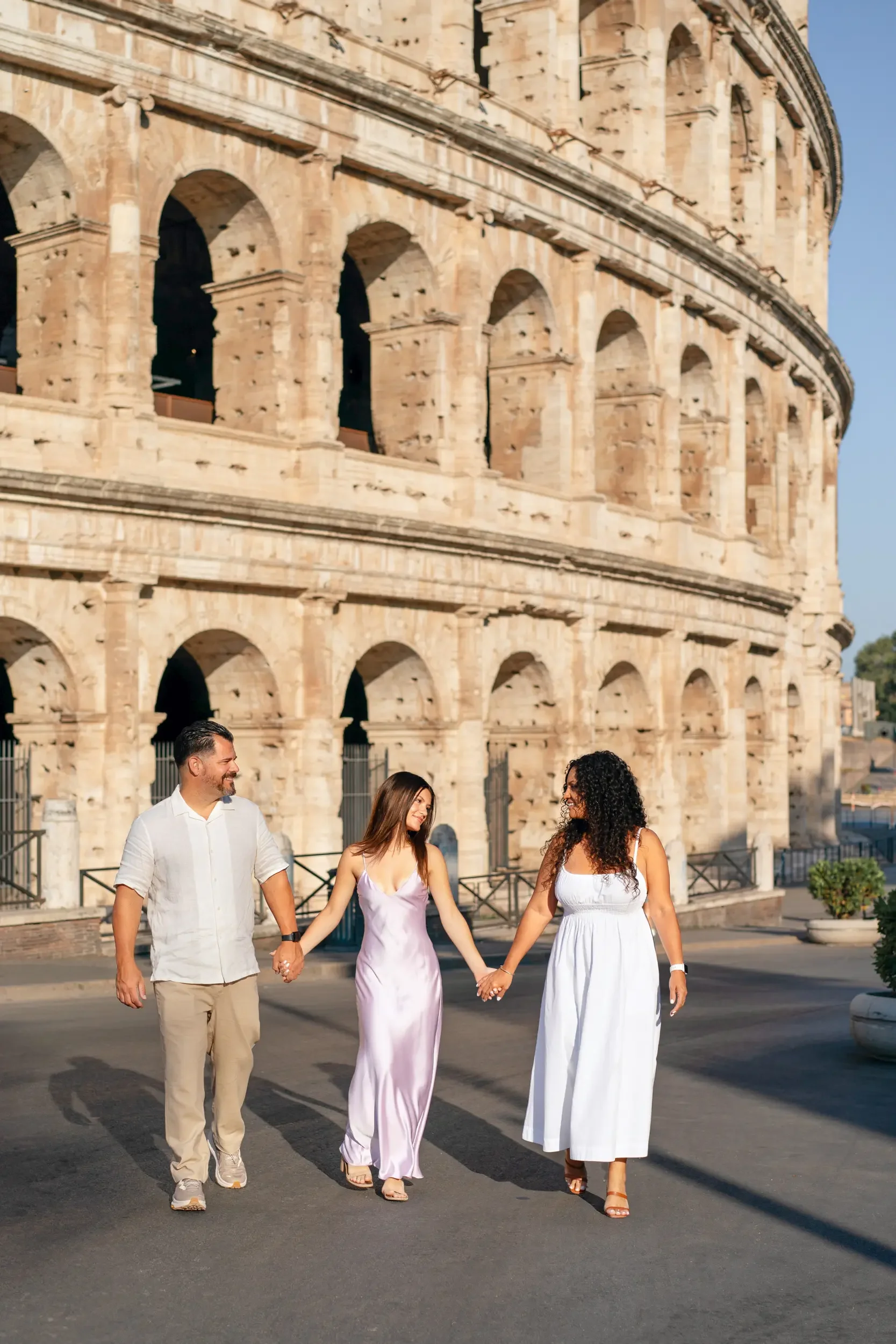 Three people, two women and one man, holding hands and walking near the Colosseum in Rome, Italy, during daylight.