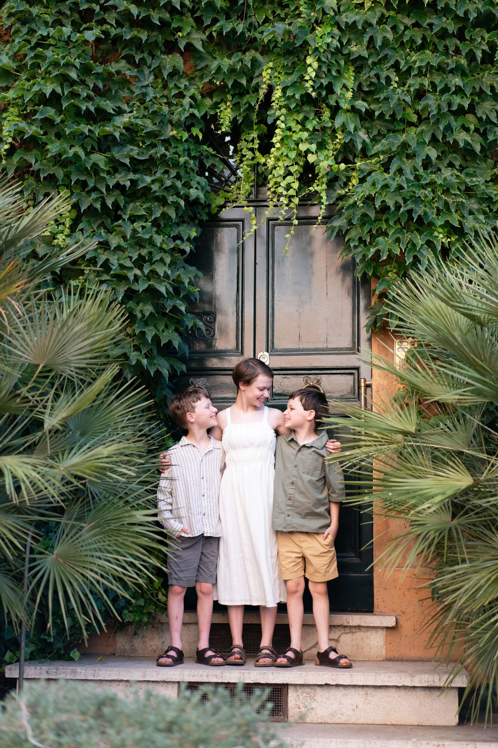 A woman and two boys standing together in front of a black gate covered with green ivy, smiling and looking at each other.