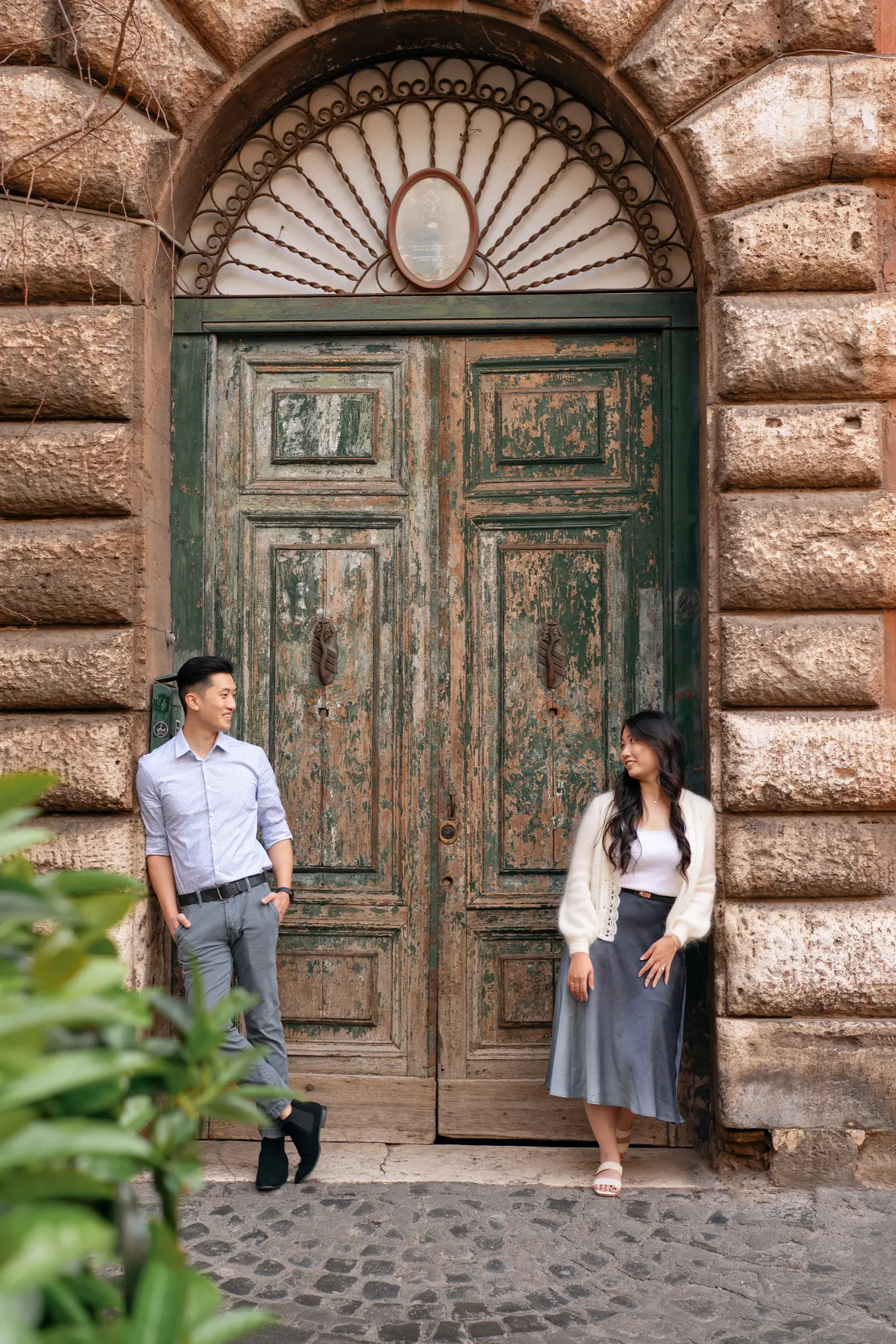 A young man and woman standing in front of a large, weathered green wooden door with stone walls on either side. The man is leaning against the wall on the left, smiling and looking at the woman. The woman is standing on the right, leaning against th