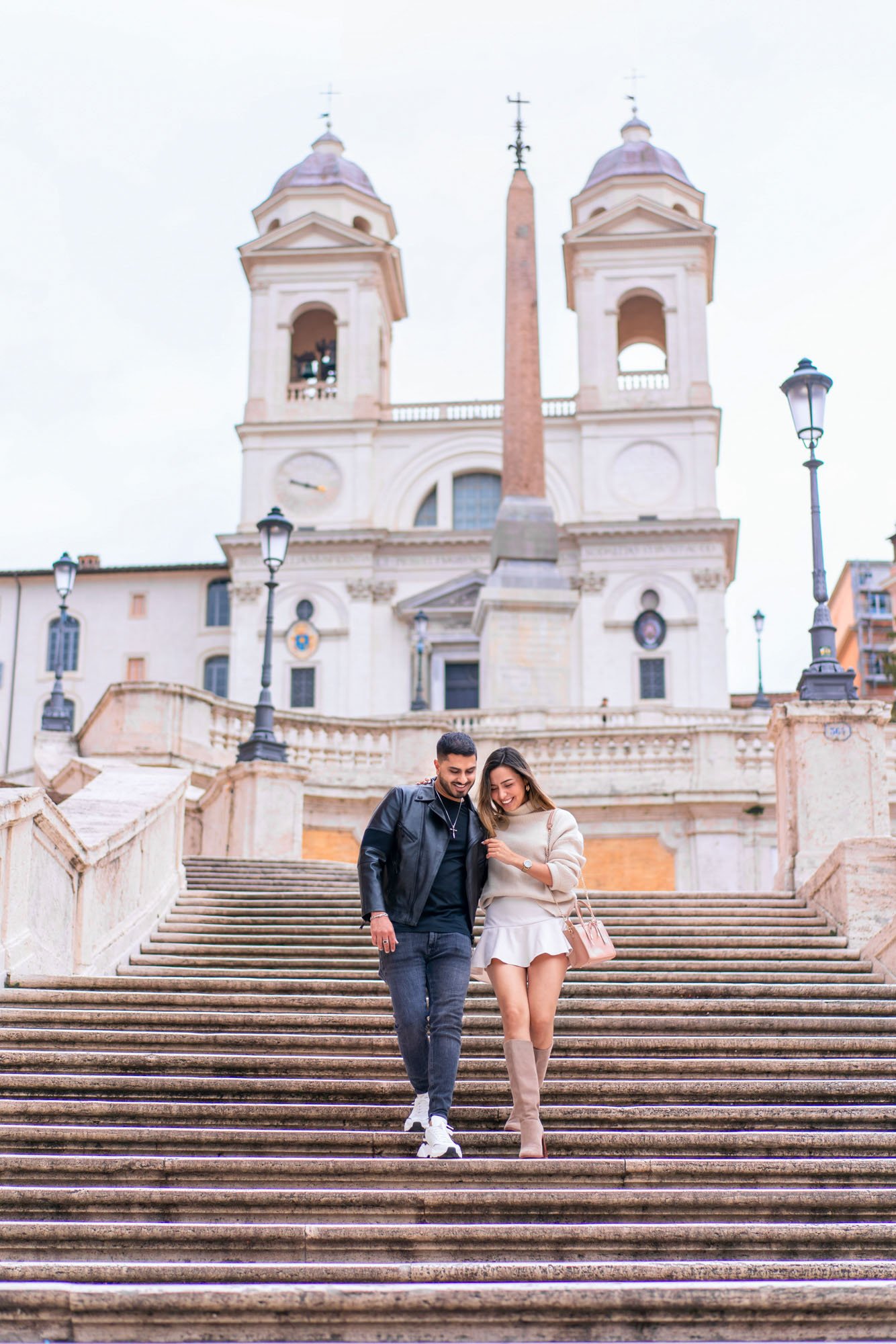 A couple walking down the stairs in front of a historic church with two towers and an obelisk in the background.