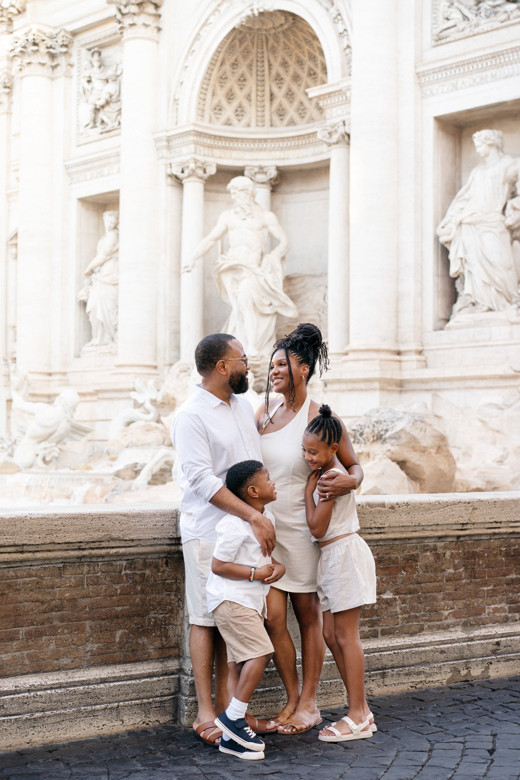 A family of four standing in front of the Trevi Fountain. The family includes a man, woman, and two children, all smiling and embracing each other. The background features ornate white sculptures and architecture of the fountain.