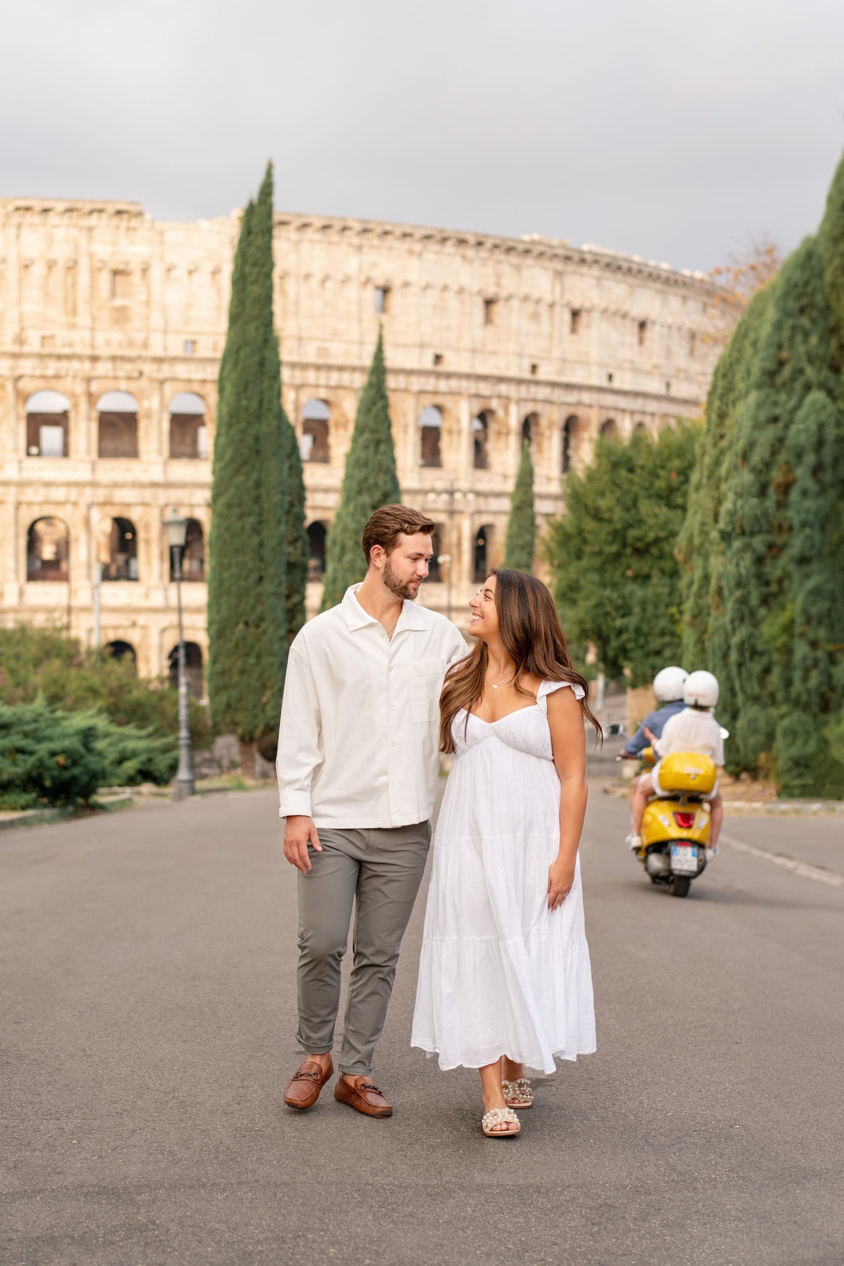 A man and a woman walking and smiling near the Colosseum in Rome, Italy, with a yellow scooter and two riders in helmets passing by in the background.