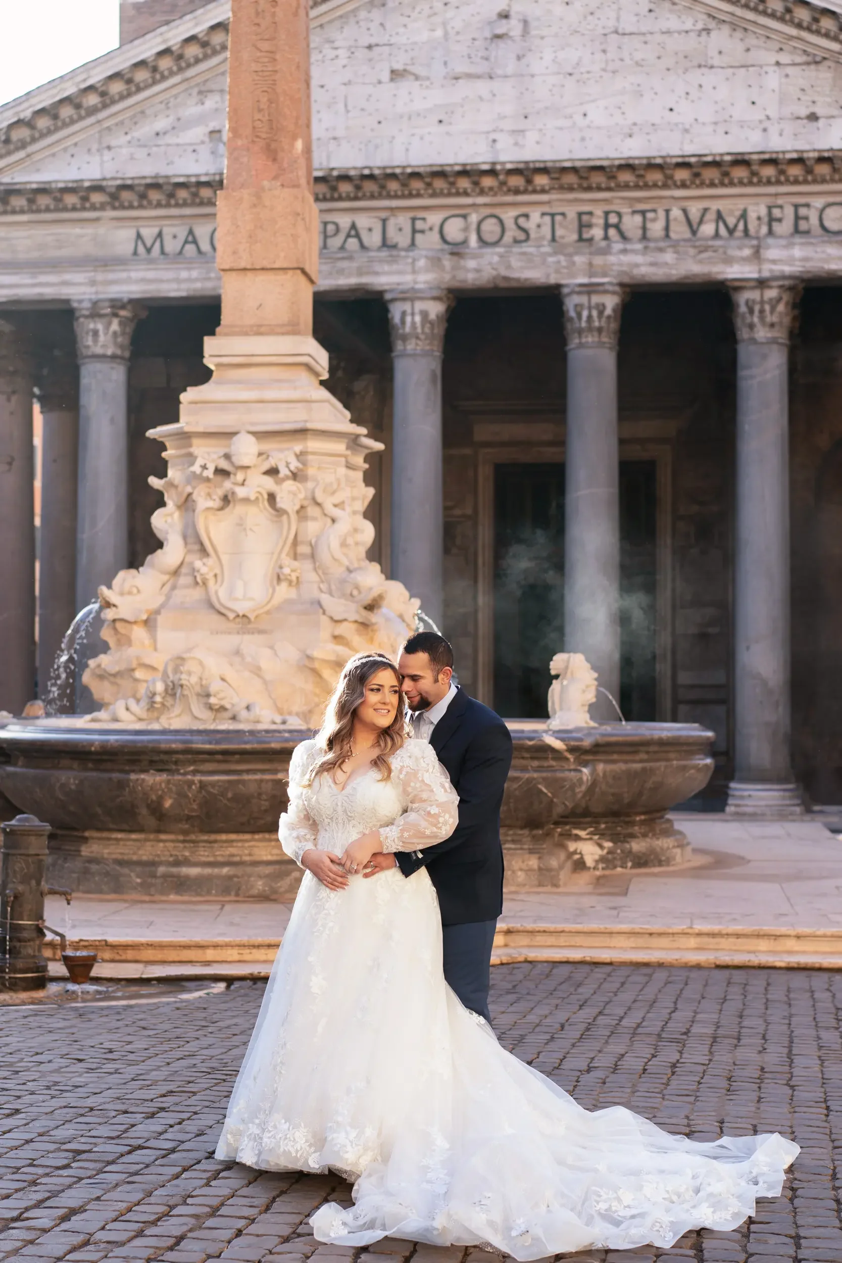 A bride and groom standing close together in front of a historic fountain and building, dressed in wedding attire, with the bride in a white lace gown and the groom in a dark suit, during golden hour.