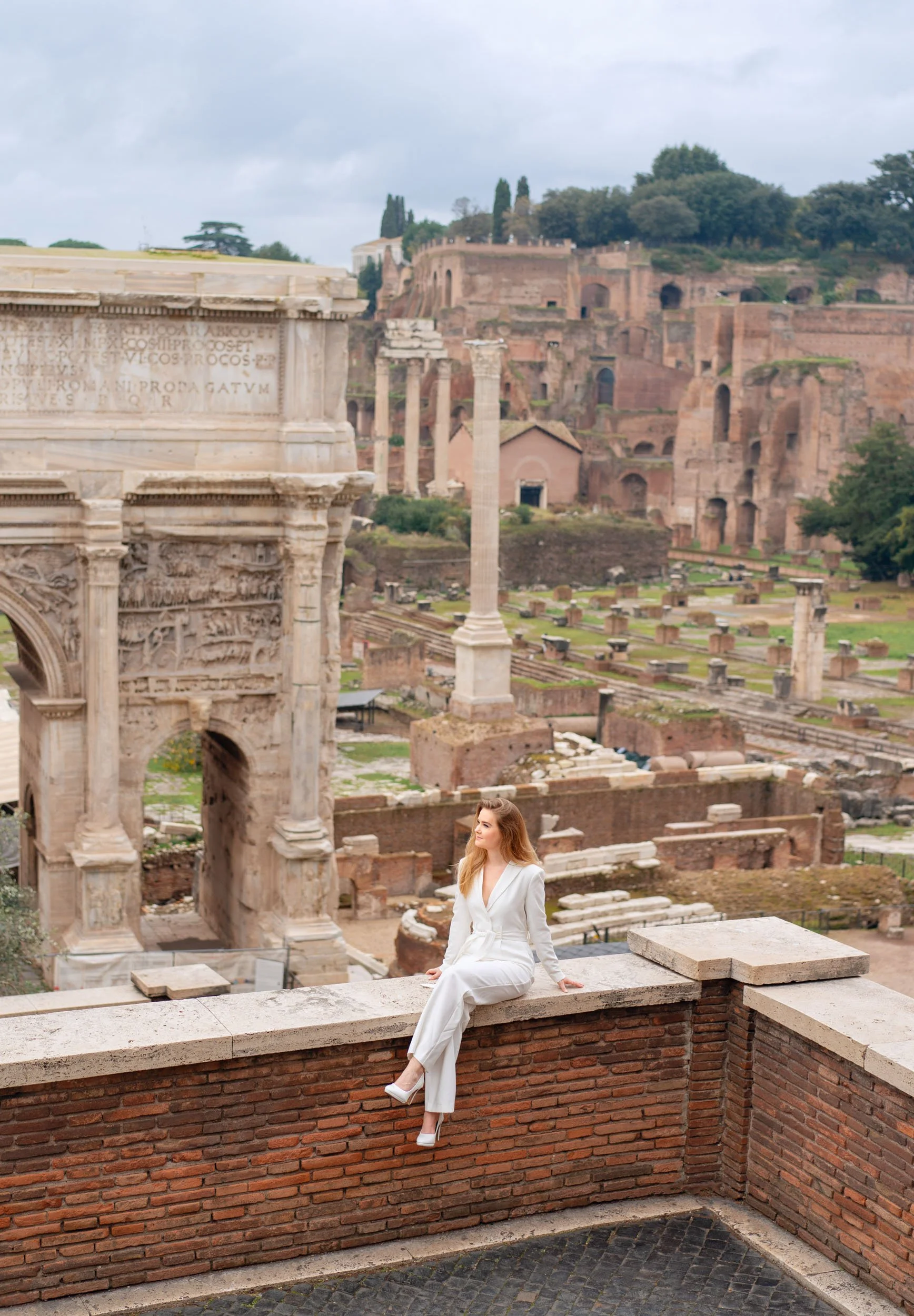 A woman in a white suit sitting on a brick wall overlooking Ancient Roman ruins, including columns and arches, with ruins and a hillside in the background.