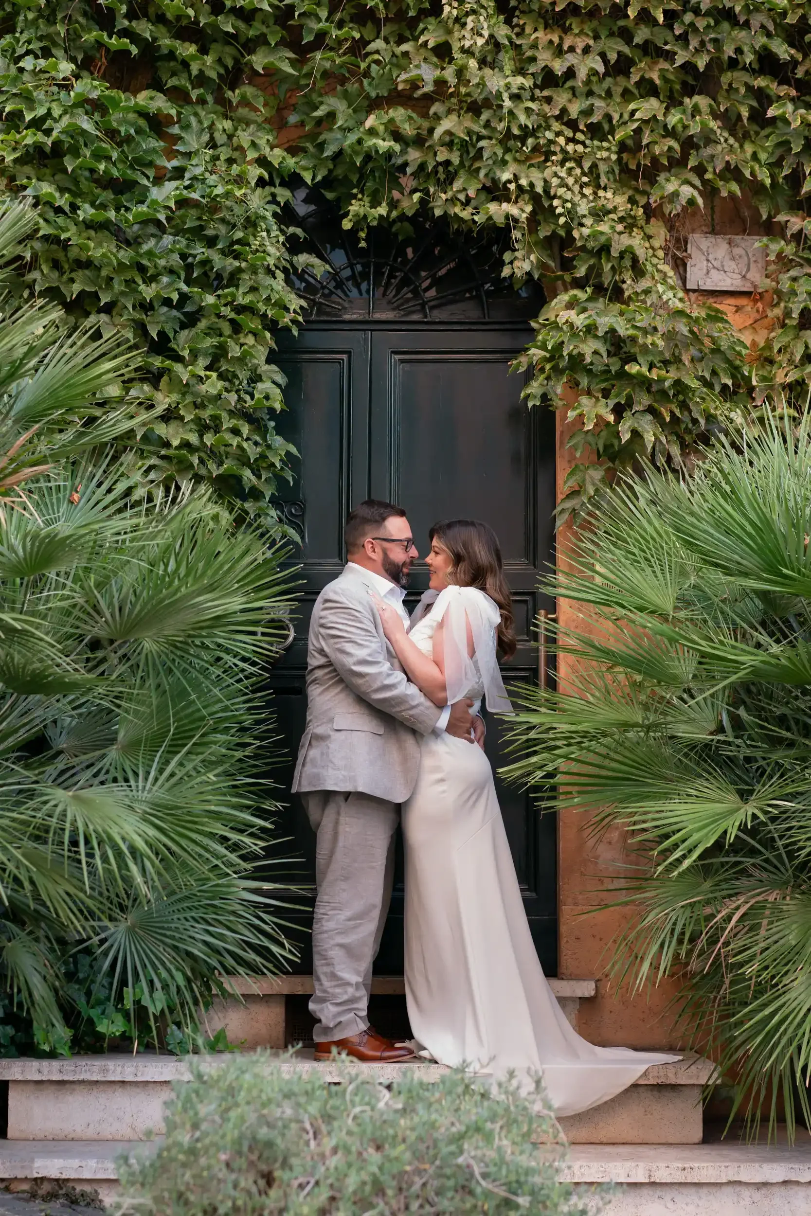 A couple in wedding attire standing close together in front of a dark green door, surrounded by lush green plants and ivy, with the woman in a white dress and the man in a light gray suit.