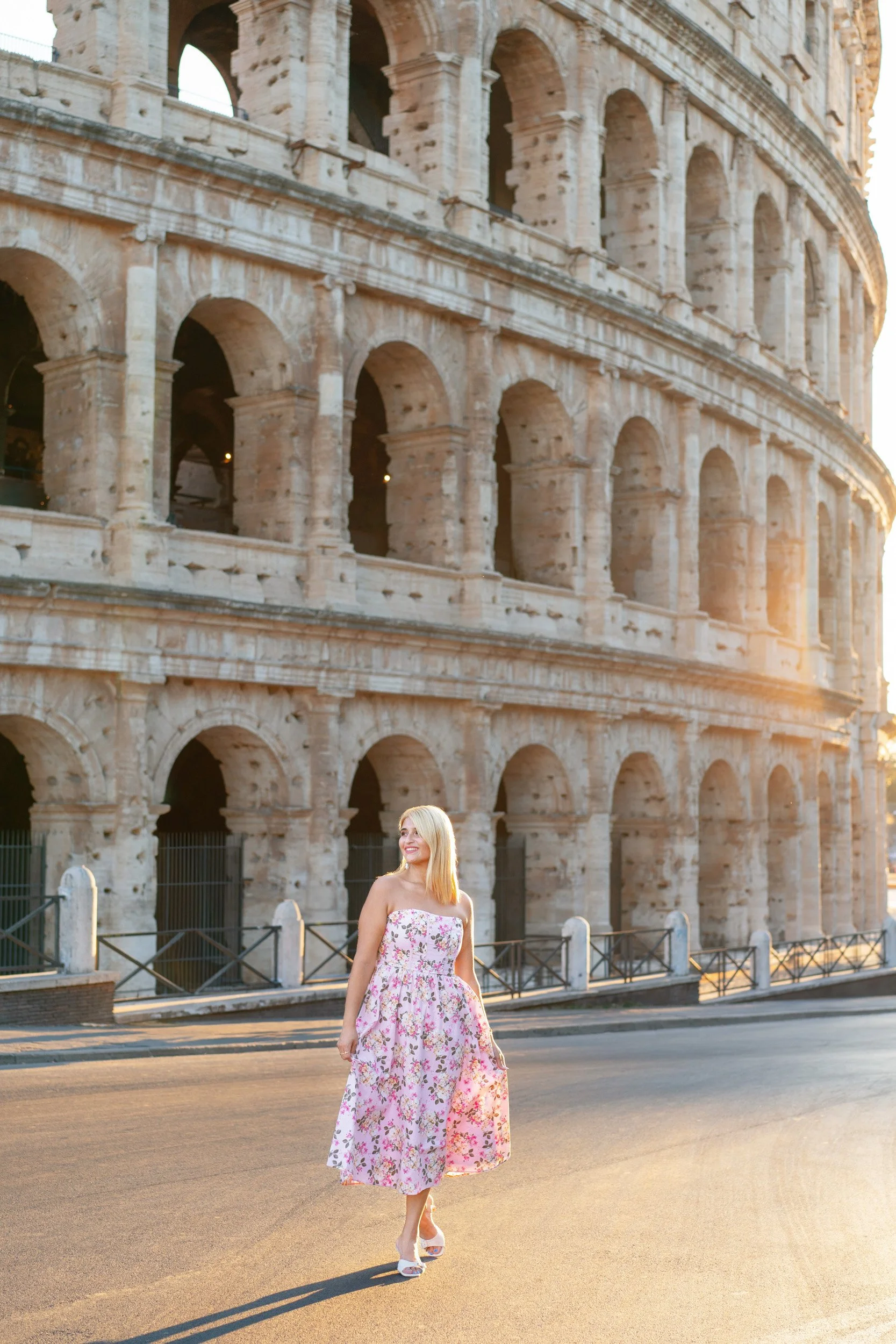 A woman in a pink floral dress walking near the Colosseum in Rome at sunset.