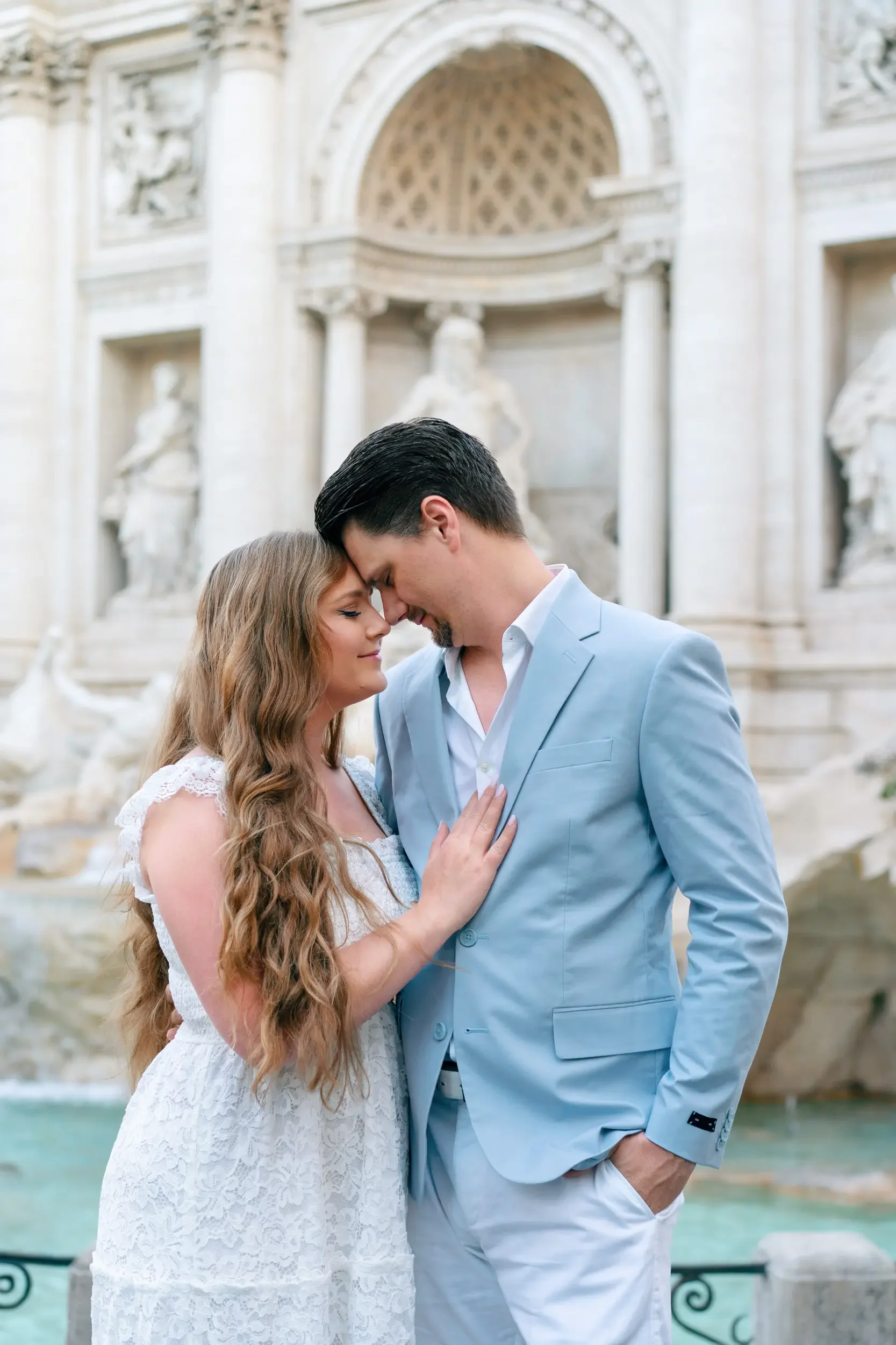 A couple sharing an intimate moment with their foreheads touching, standing in front of a historic fountain and ornate architecture.