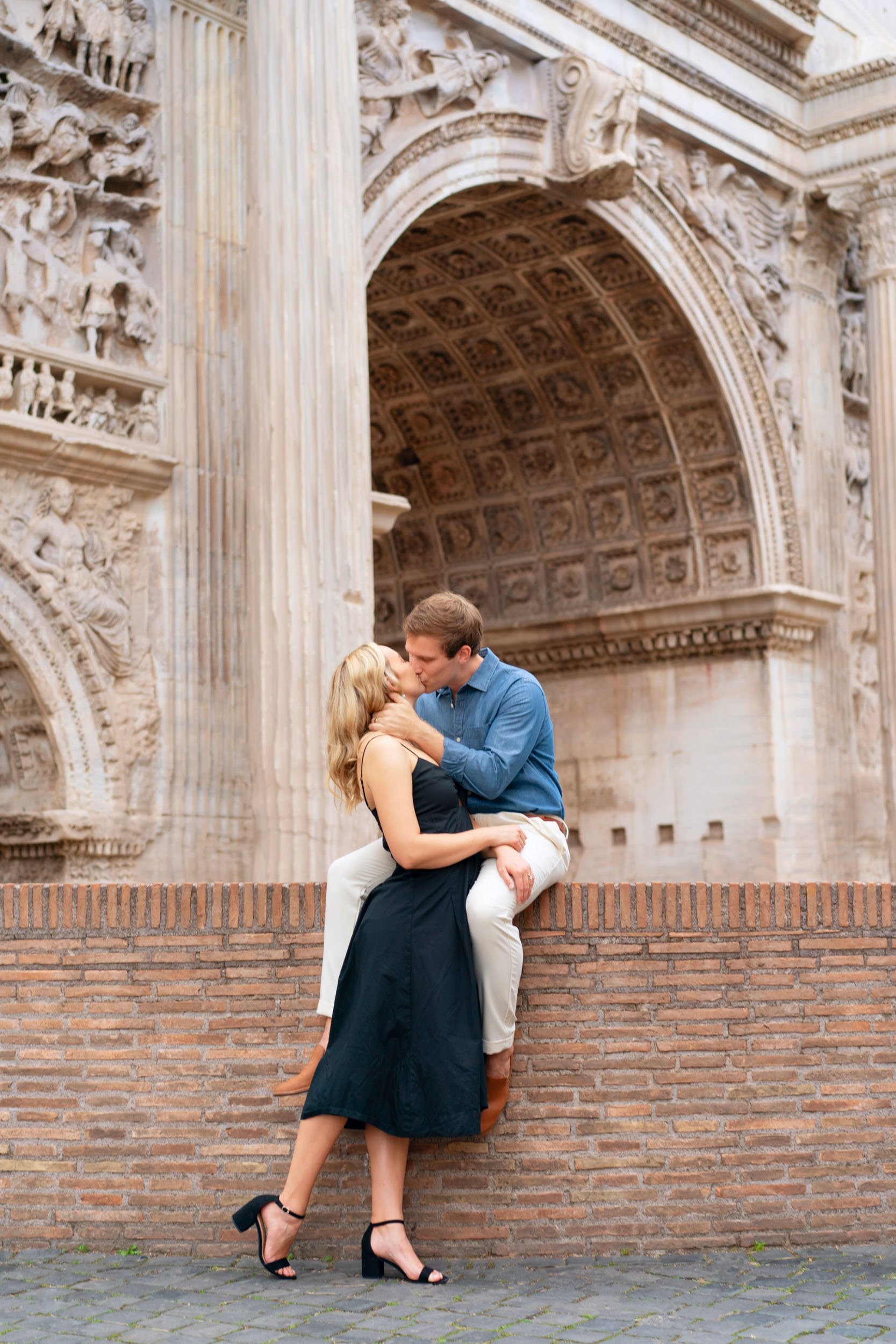 A couple kissing on a brick wall in front of an ancient, ornate stone archway with intricate carvings.