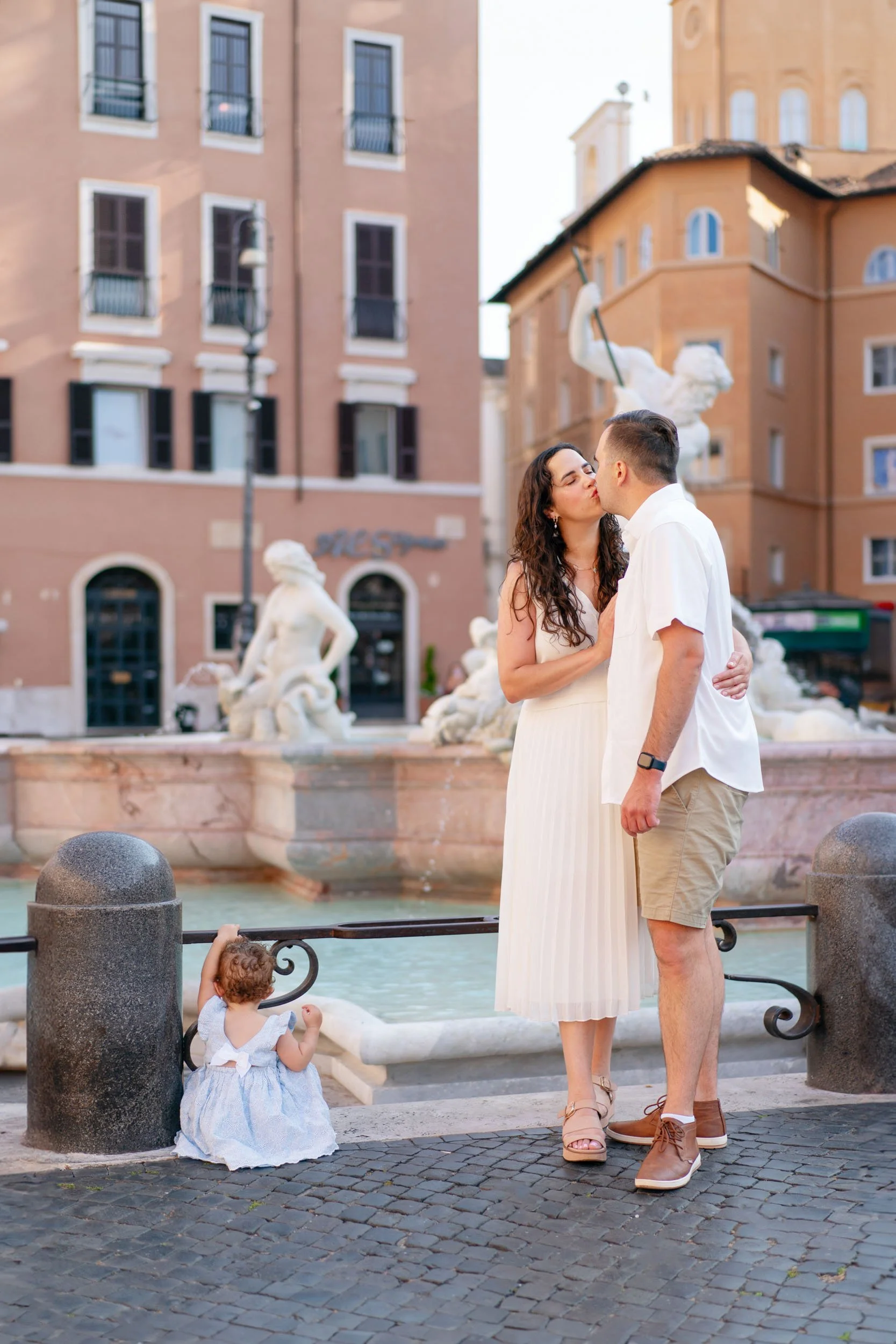 A couple kissing in front of a fountain in a city square, with a toddler sitting nearby and buildings in the background.
