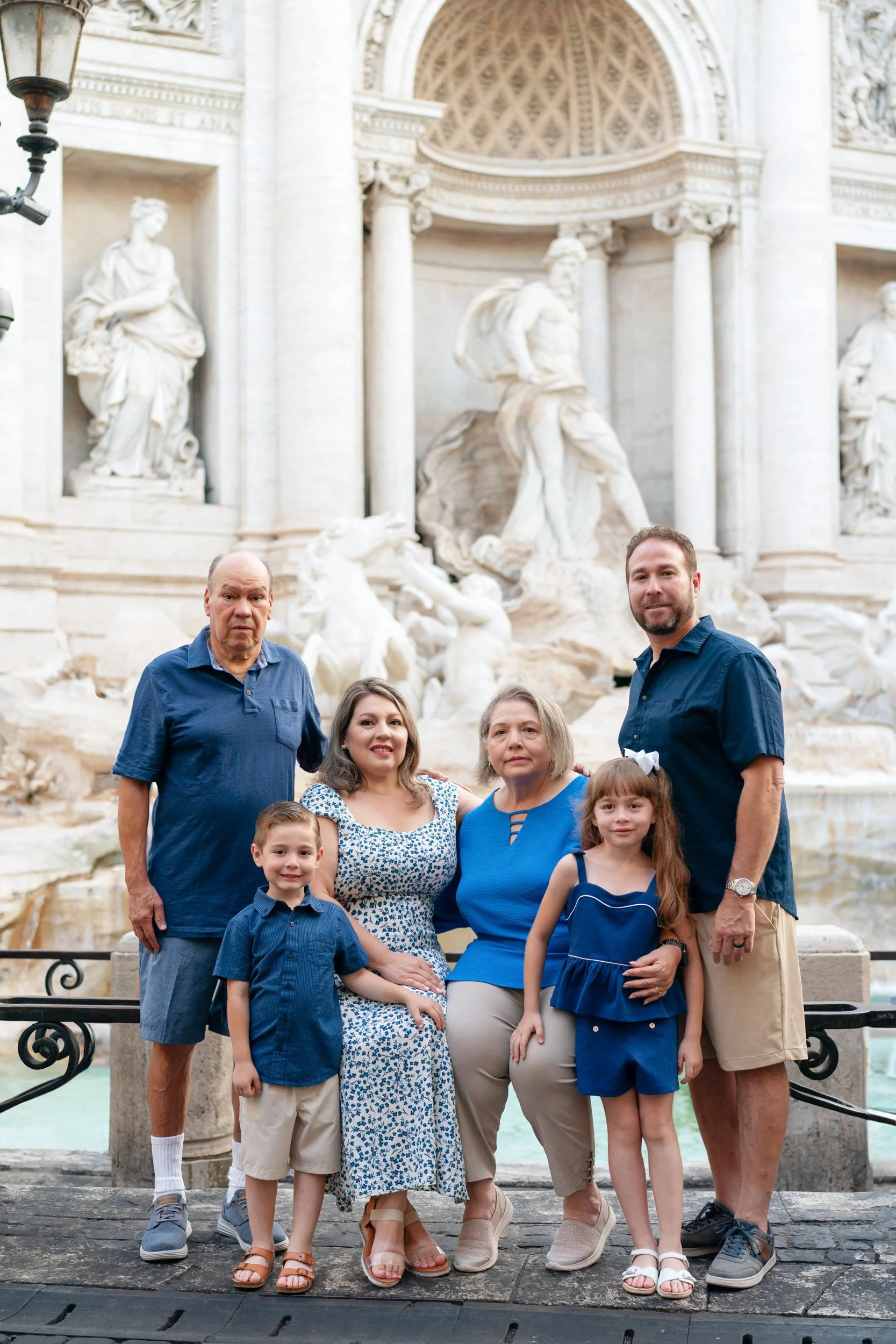 A family of six posing in front of the Trevi Fountain in Rome, Italy. The family includes two older adults, a man and a woman, and four children, standing on a stone platform with the ornate fountain and statues in the background.