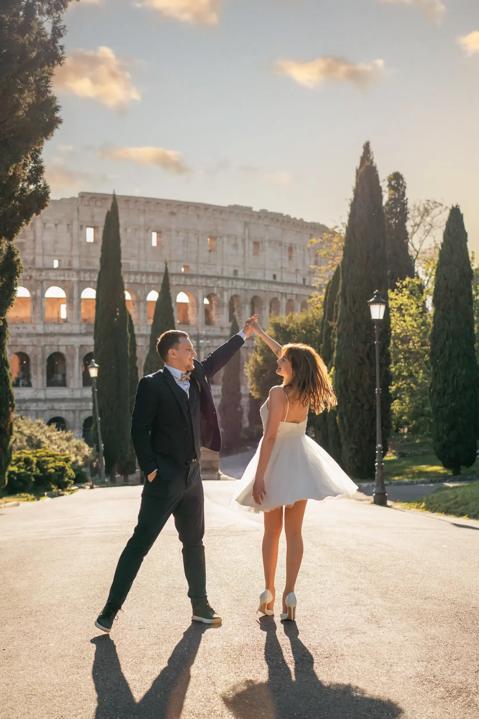 A man and woman dancing together on an empty street during sunset with the Roman Colosseum in the background, surrounded by tall cypress trees.