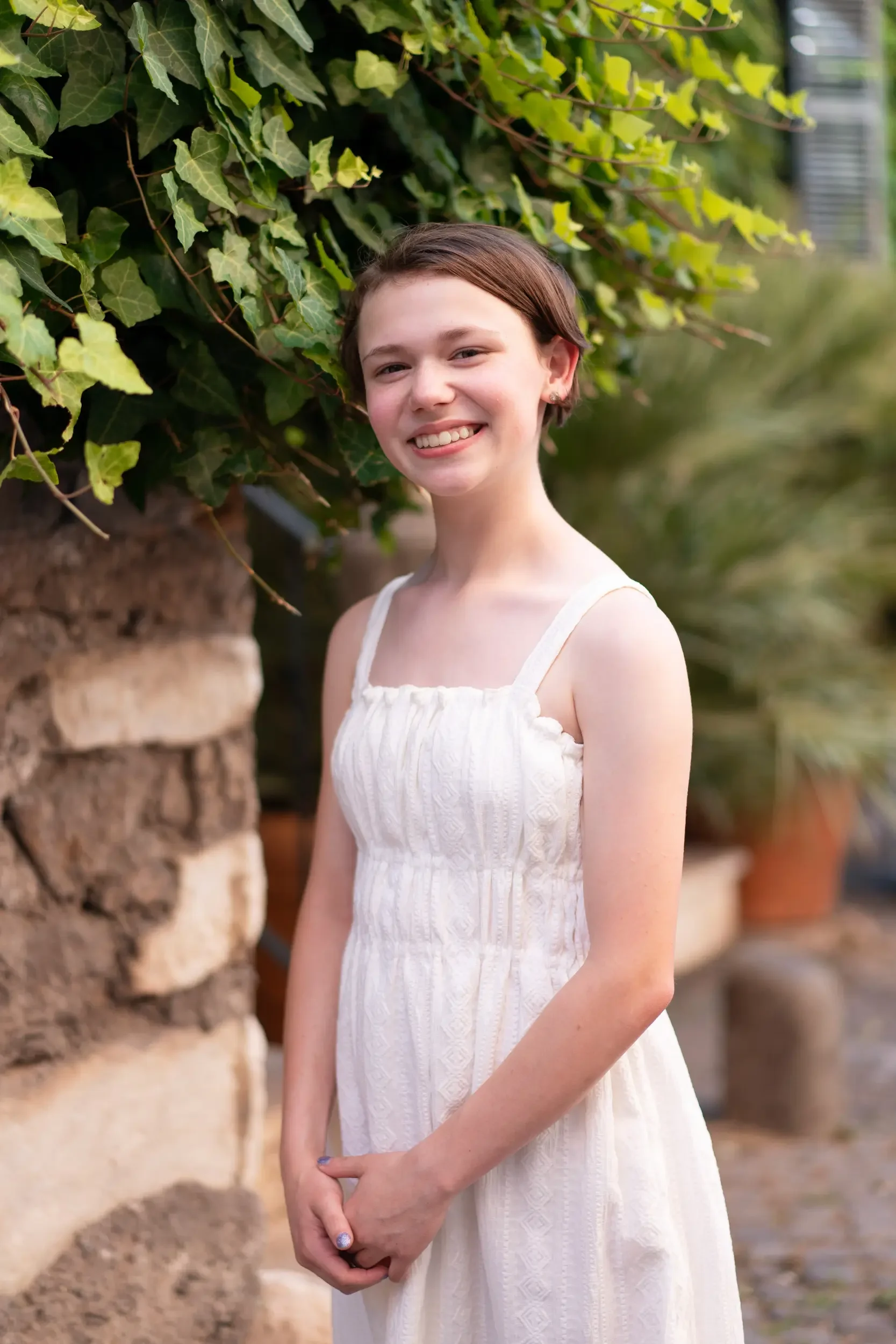 A young girl in a white dress smiling outdoors in front of green leaves and a stone wall.