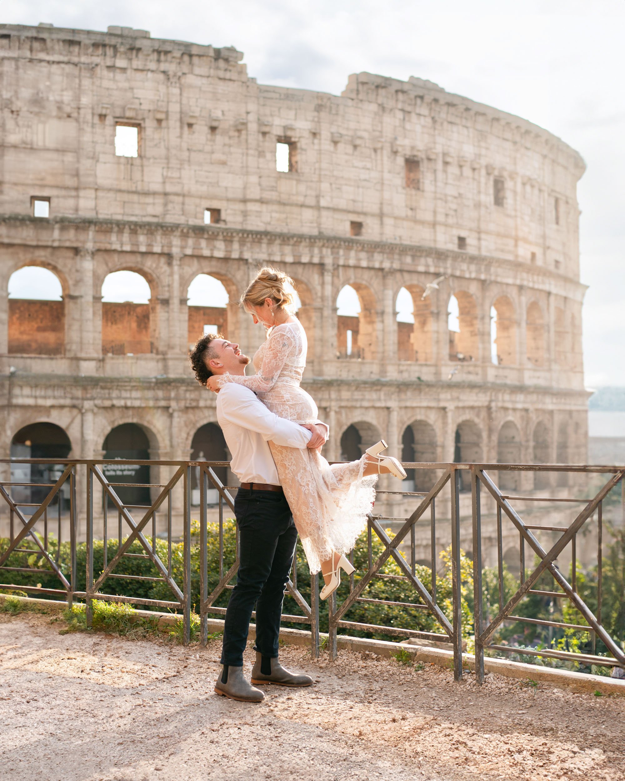 A man lifting a woman in front of the Colosseum in Rome, Italy. The woman is pregnant and wearing a lace dress; the man is smiling and wearing a white shirt and dark pants.