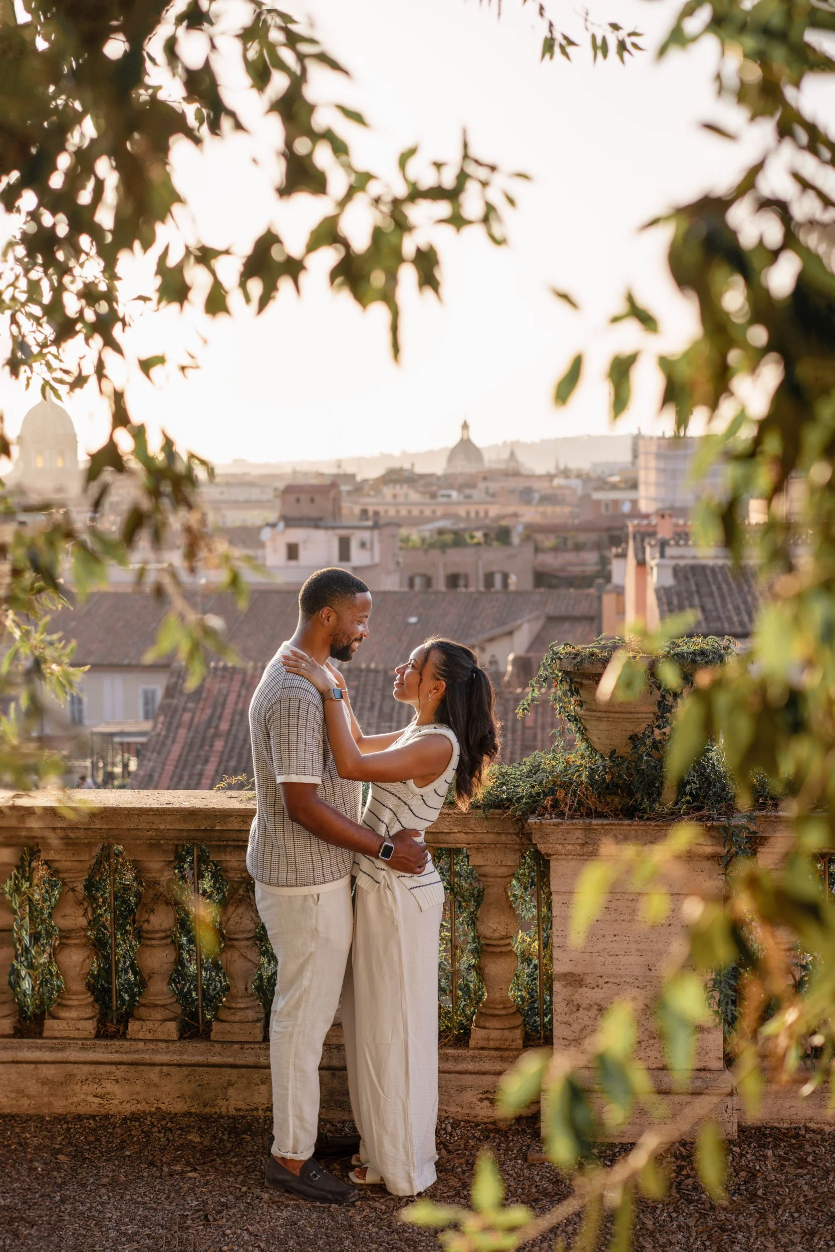 A couple standing close together on a rooftop terrace, embracing and smiling at each other with city rooftops and domed buildings in the background during sunset.