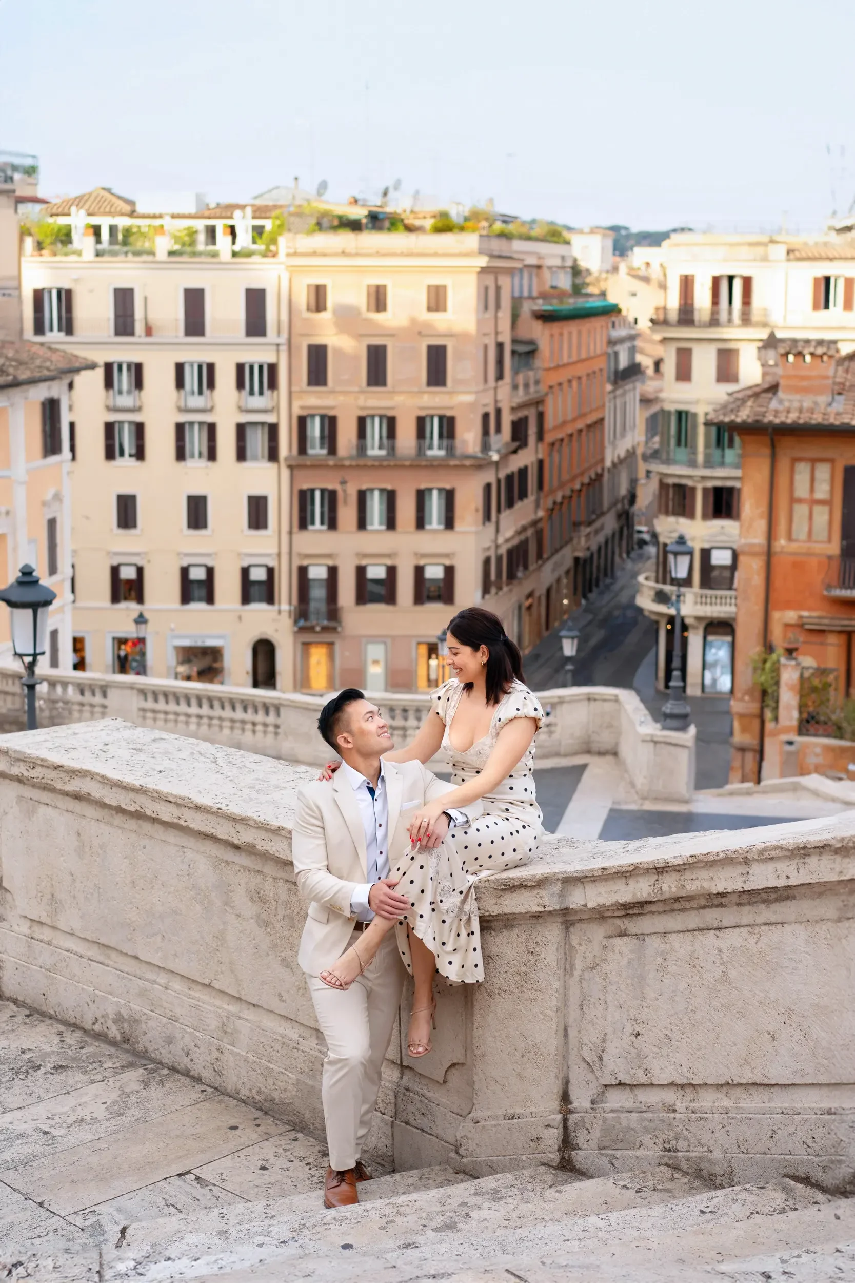 A couple on a stone staircase in a European city with colorful buildings in the background, smiling and looking at each other.