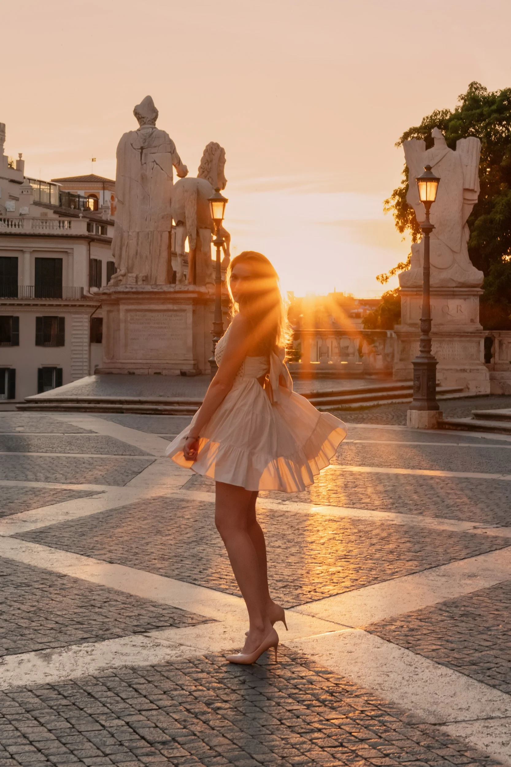 A woman in a beige dress and high heels standing in a city square during sunset, with statues and lamp posts in the background.