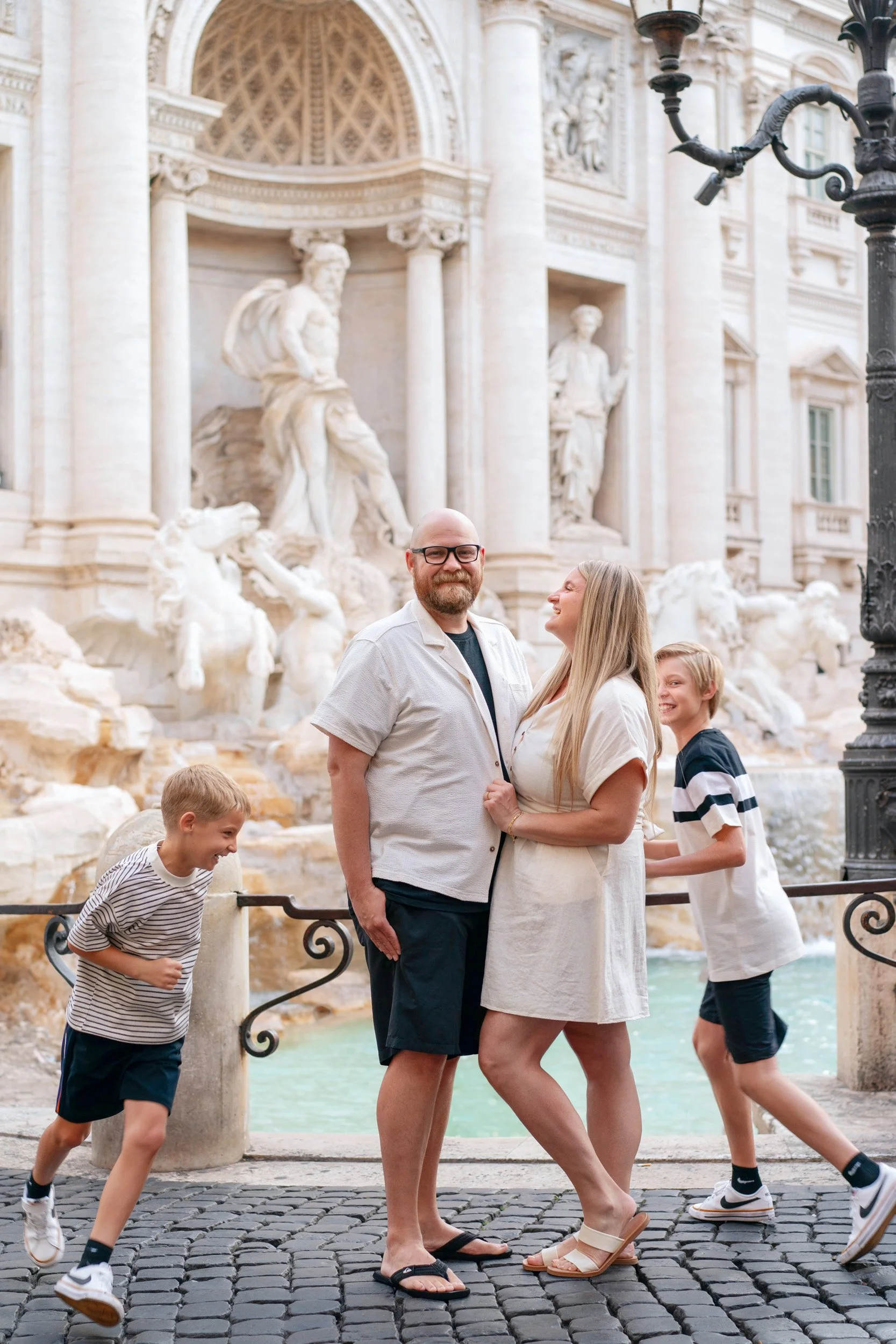 A family of four standing in front of the Trevi Fountain in Rome, Italy, with the fountain's ornate sculptures and water feature in the background. The parents are smiling and holding each other, while the two children are playing and laughing nearby