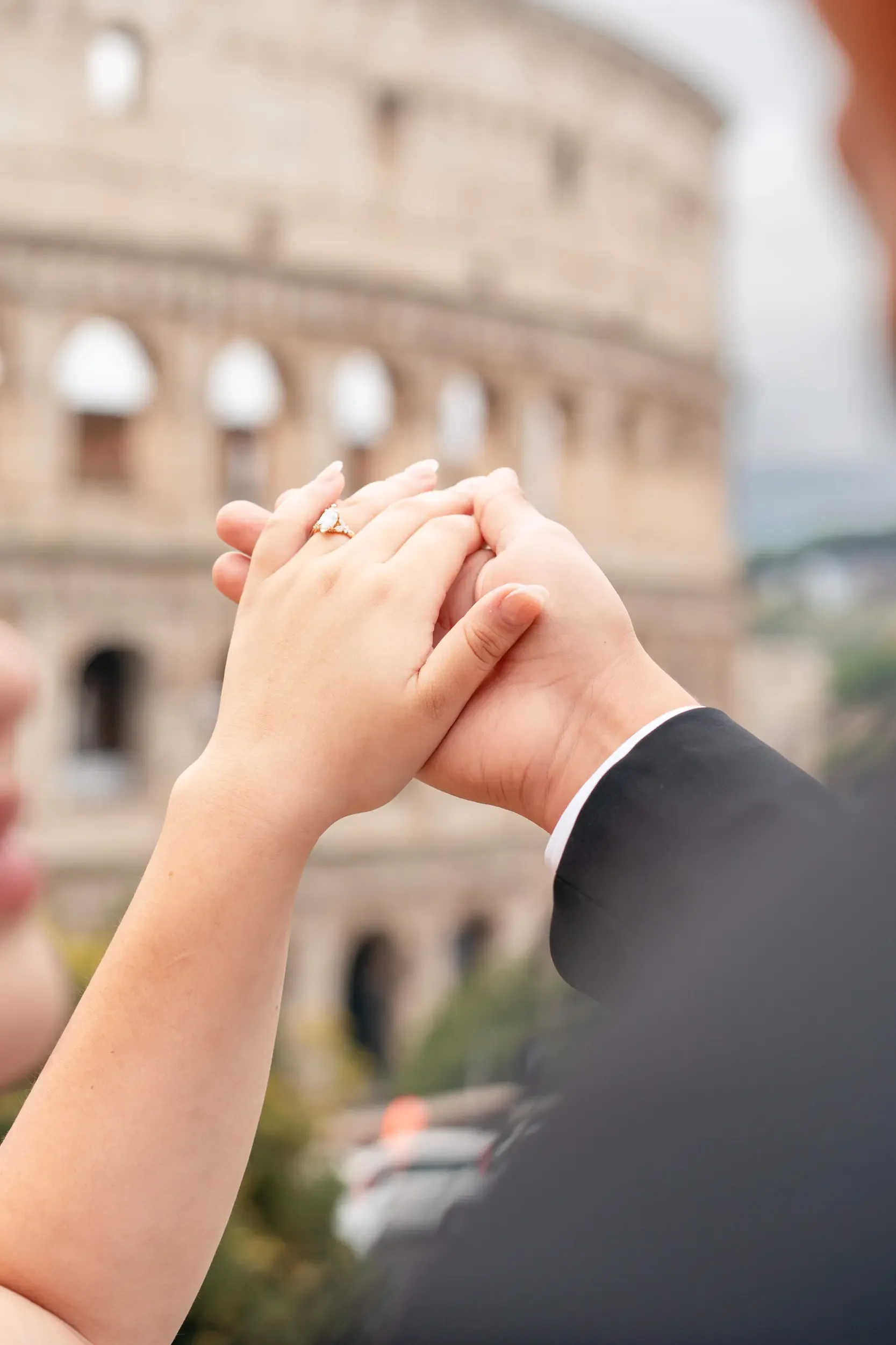 A close-up of a couple holding hands, showing a wedding ring on the woman's finger, with the Colosseum in the background.