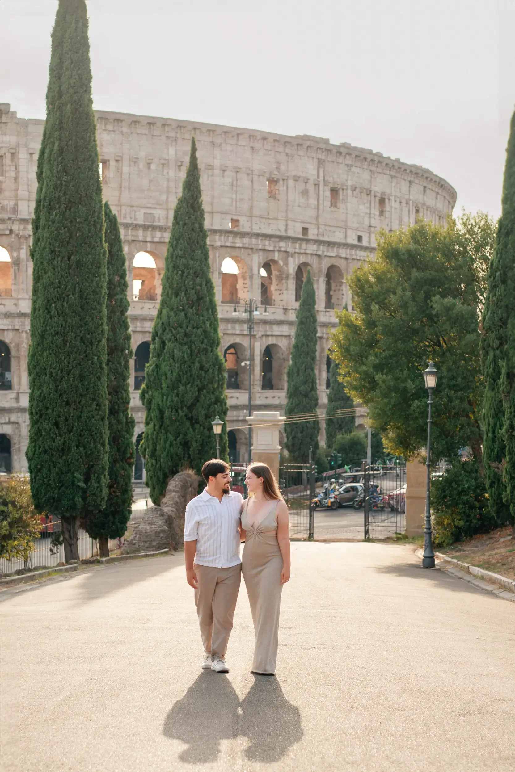 A couple walking arm in arm in front of the Colosseum in Rome, Italy, with tall cypress trees and greenery around them, during daytime.