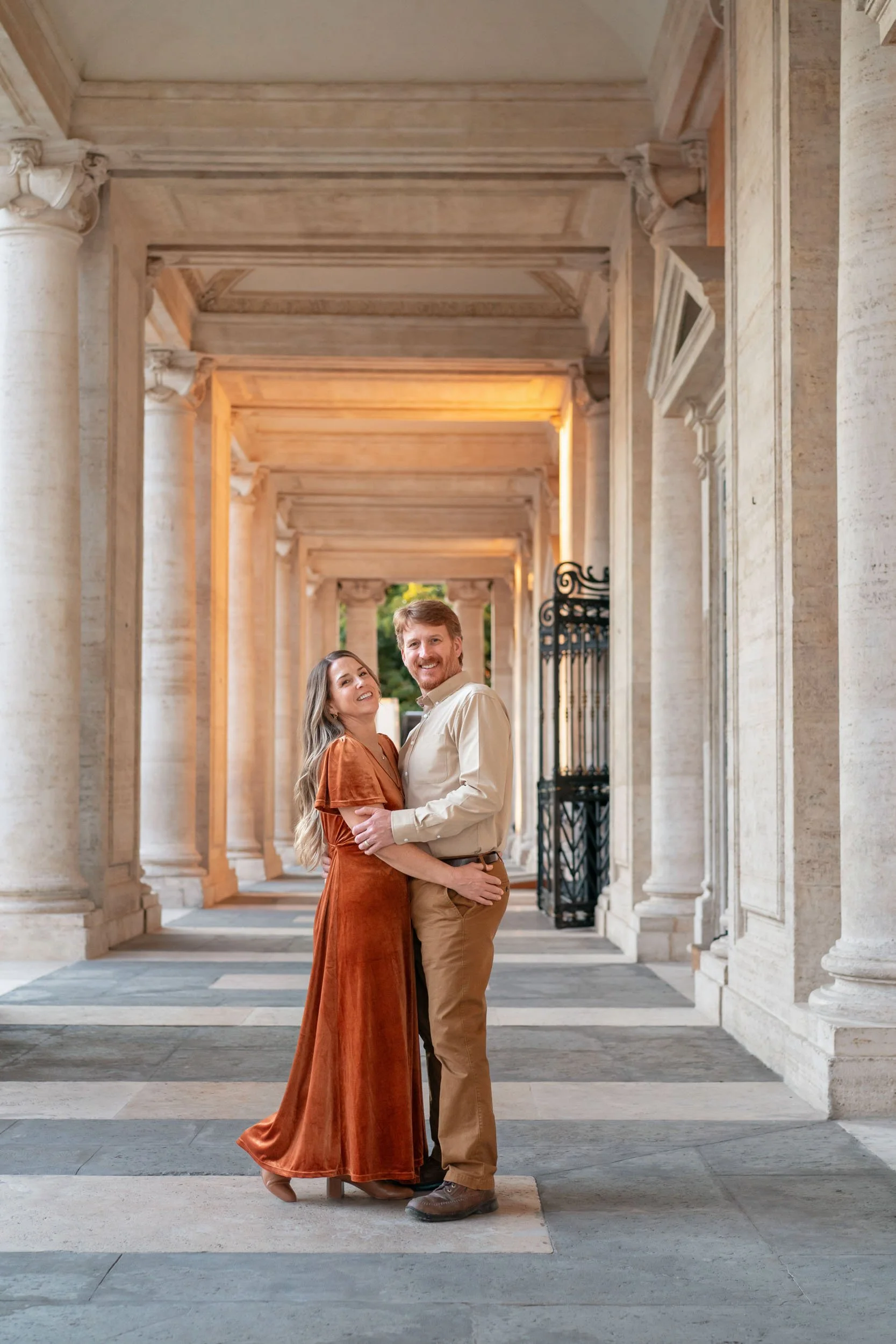 A smiling couple embracing in a colonnade with stone columns and arches, during sunset.