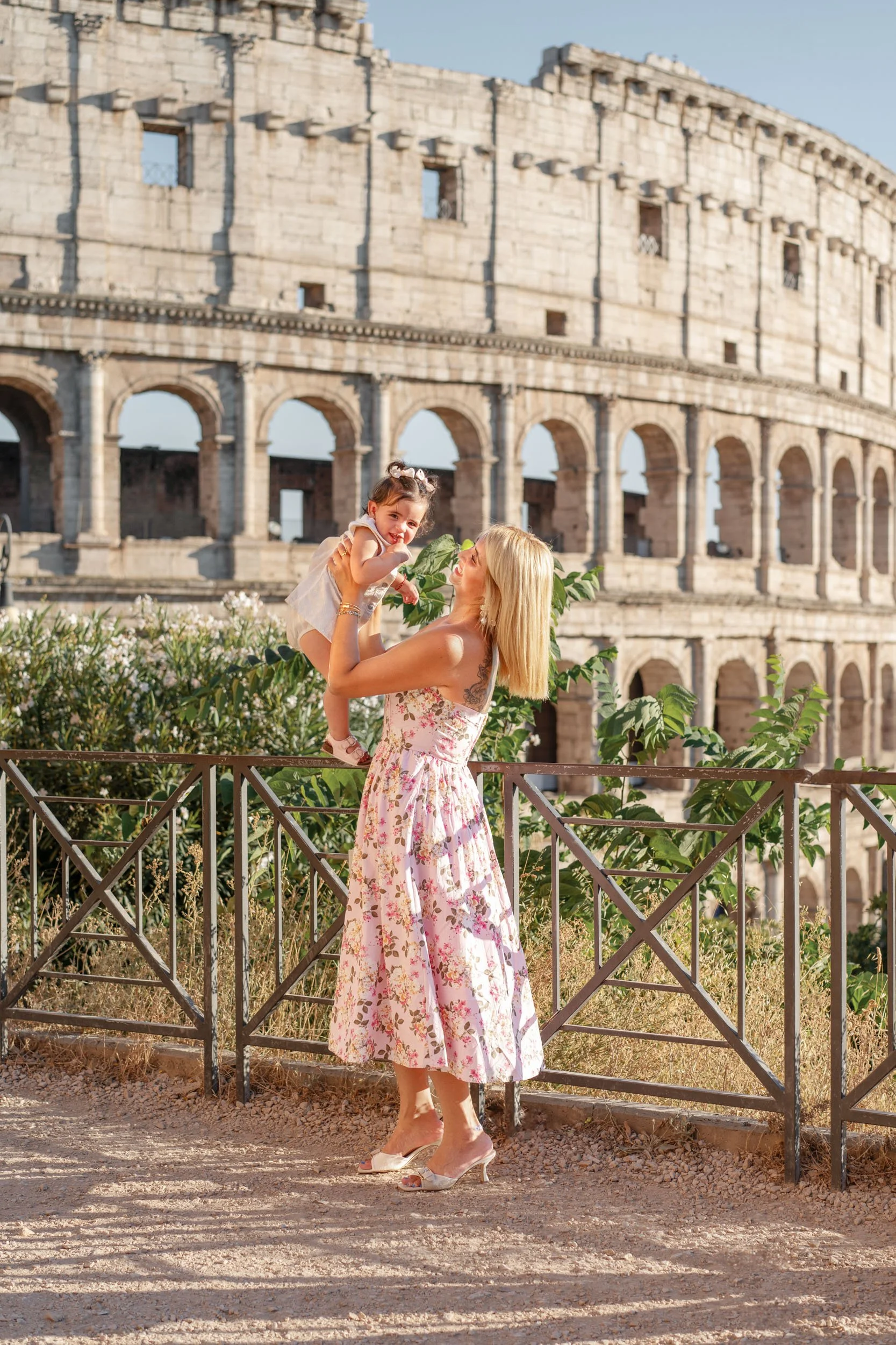 A woman in a floral dress lifting a young girl in front of the Roman Colosseum, with green bushes and a metal fence in the foreground.