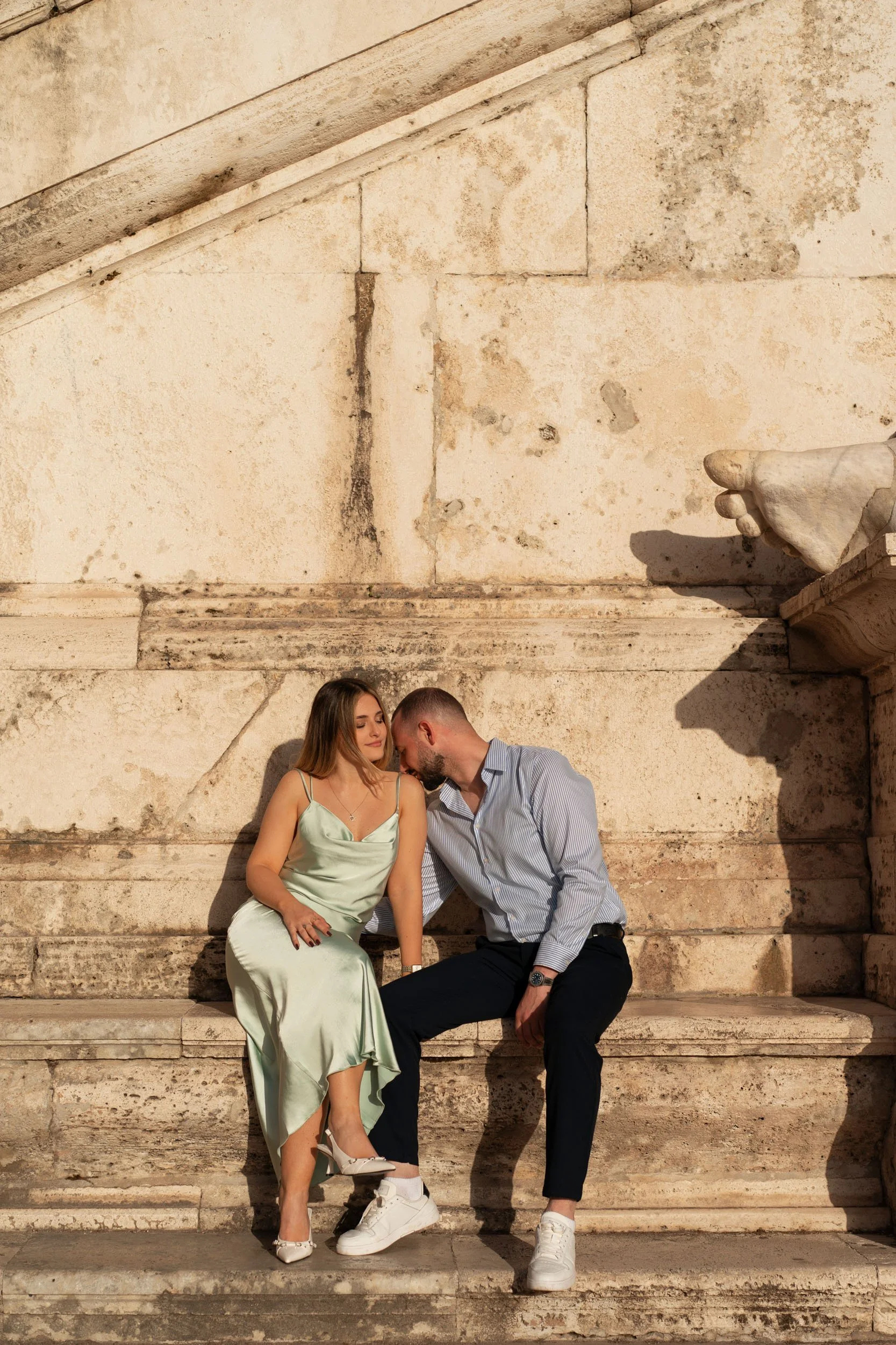 A young woman in a satin green dress and a young man in a light blue shirt and black pants sitting closely on stone steps, with their heads touching, against a weathered stone wall.