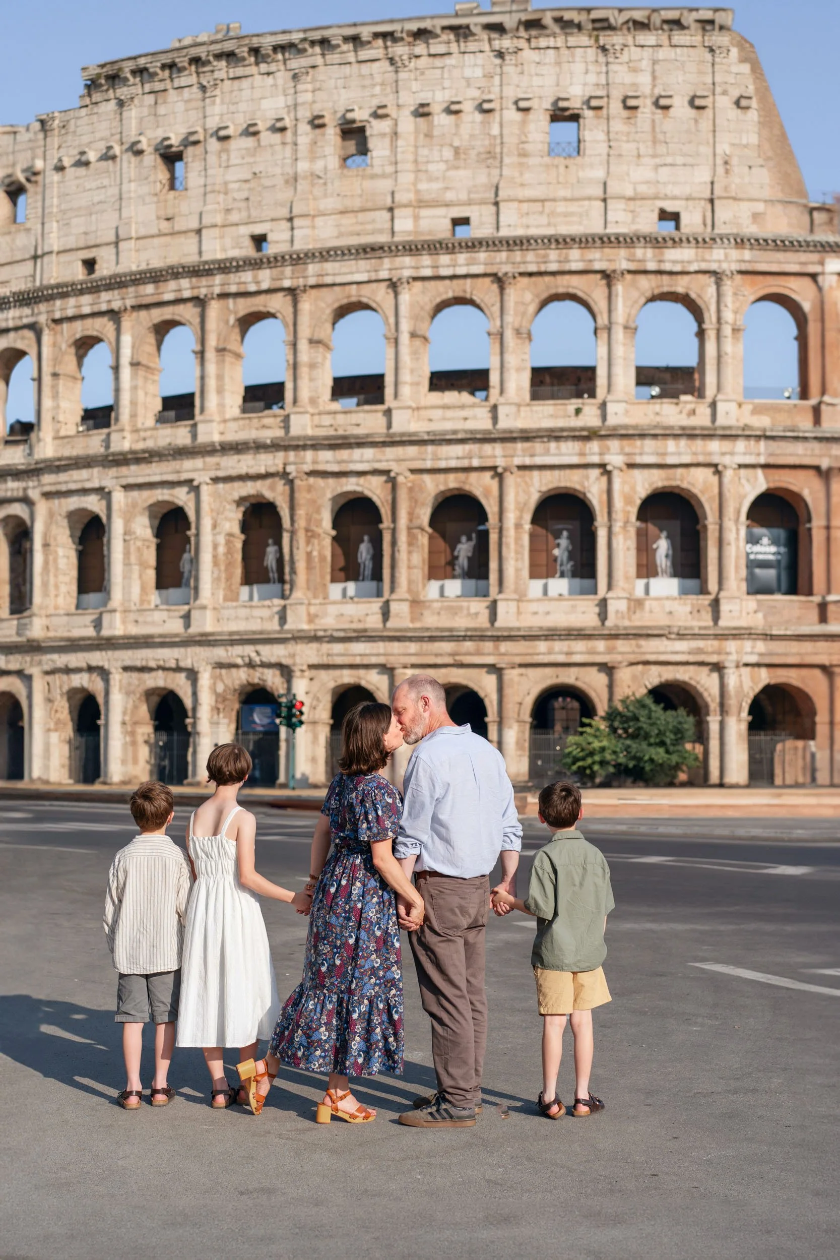 A couple kissing in front of the Colosseum with three children holding hands nearby.