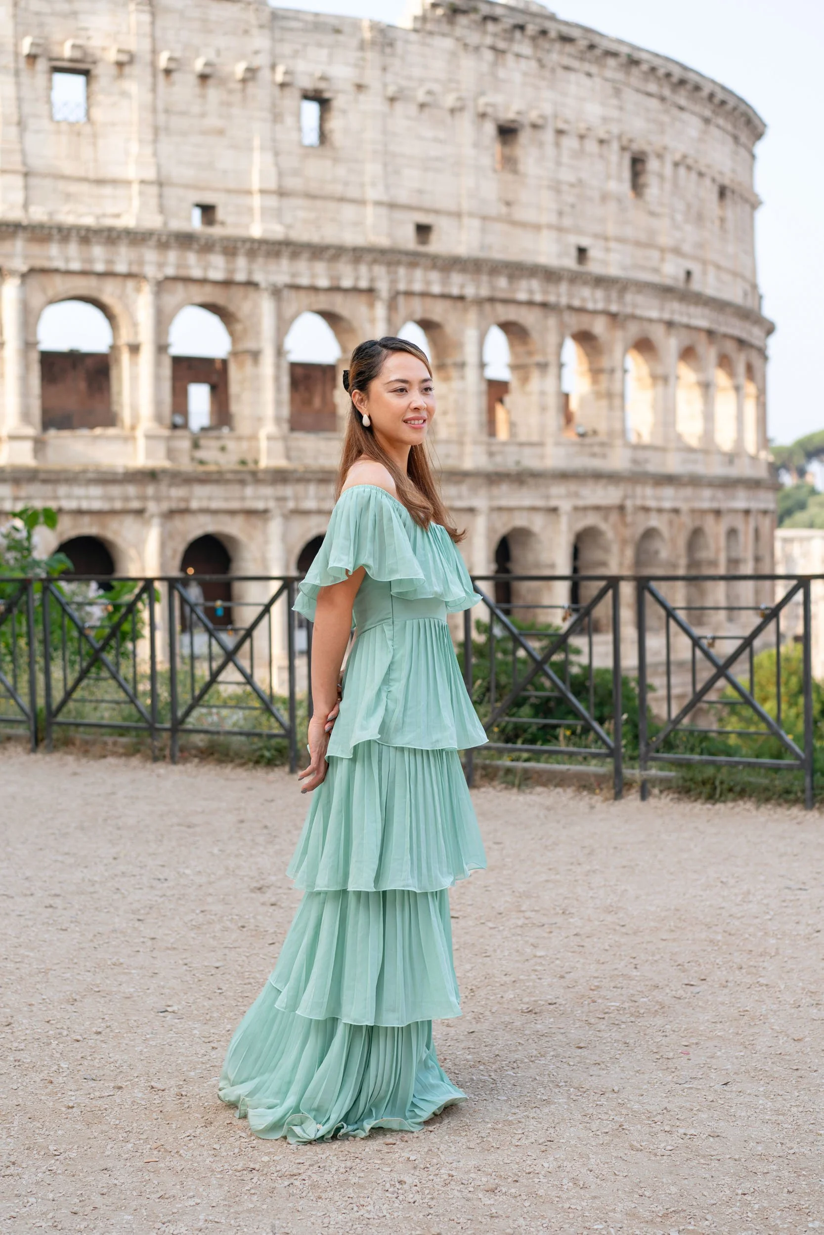 A woman in a flowing, light green dress standing outdoors in front of the Colosseum in Rome, Italy.