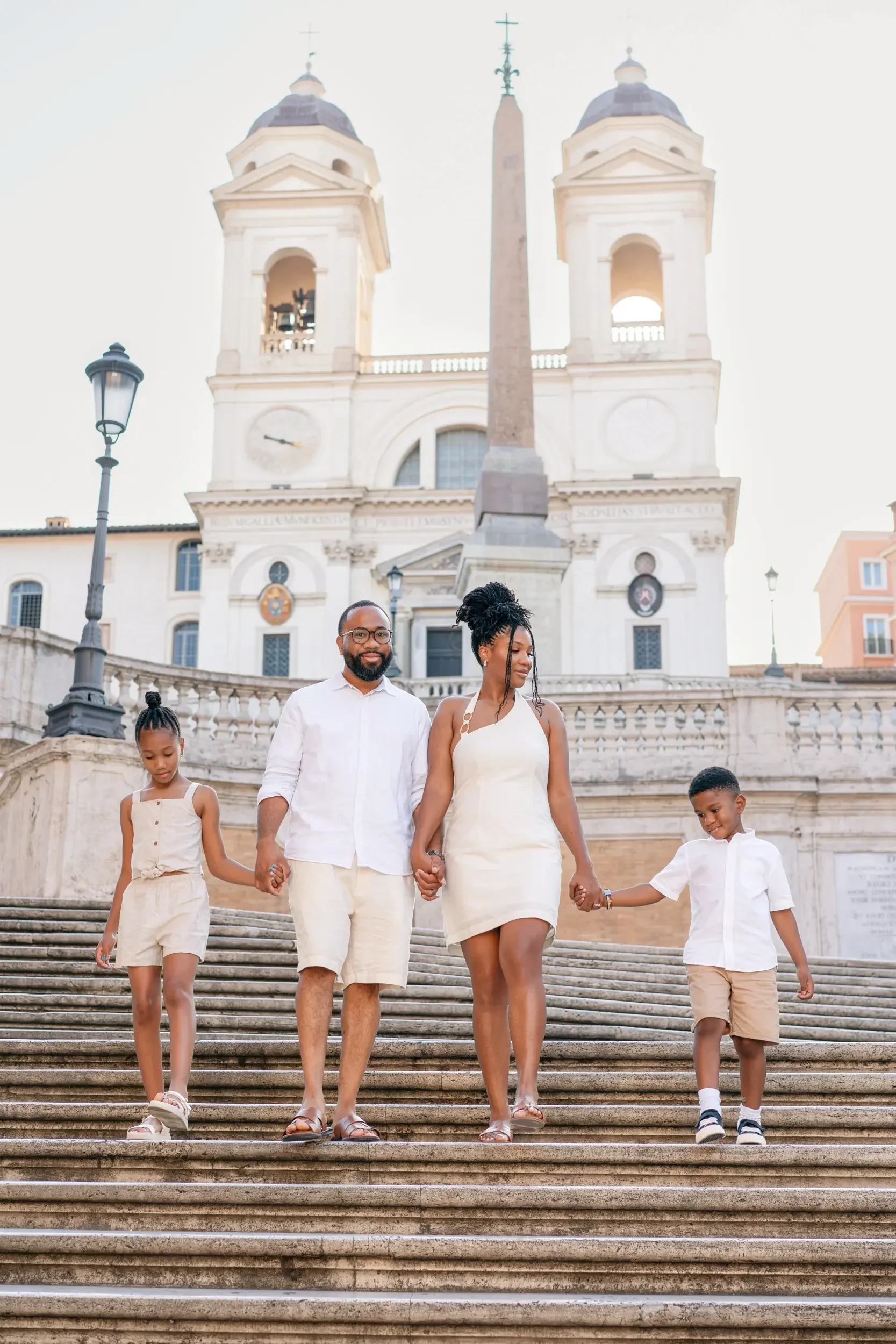 A multicultural family walking down stairs in front of a historic church, holding hands and smiling, dressed in light-colored summer clothing.