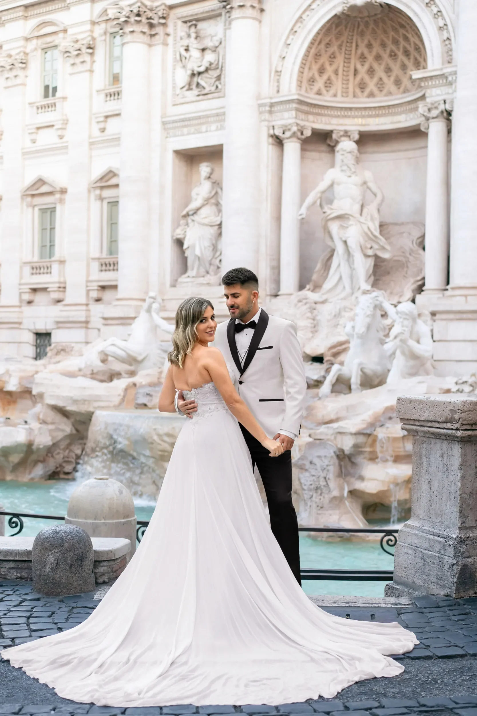 A bride and groom in wedding attire posing in front of the Trevi Fountain in Rome, Italy.