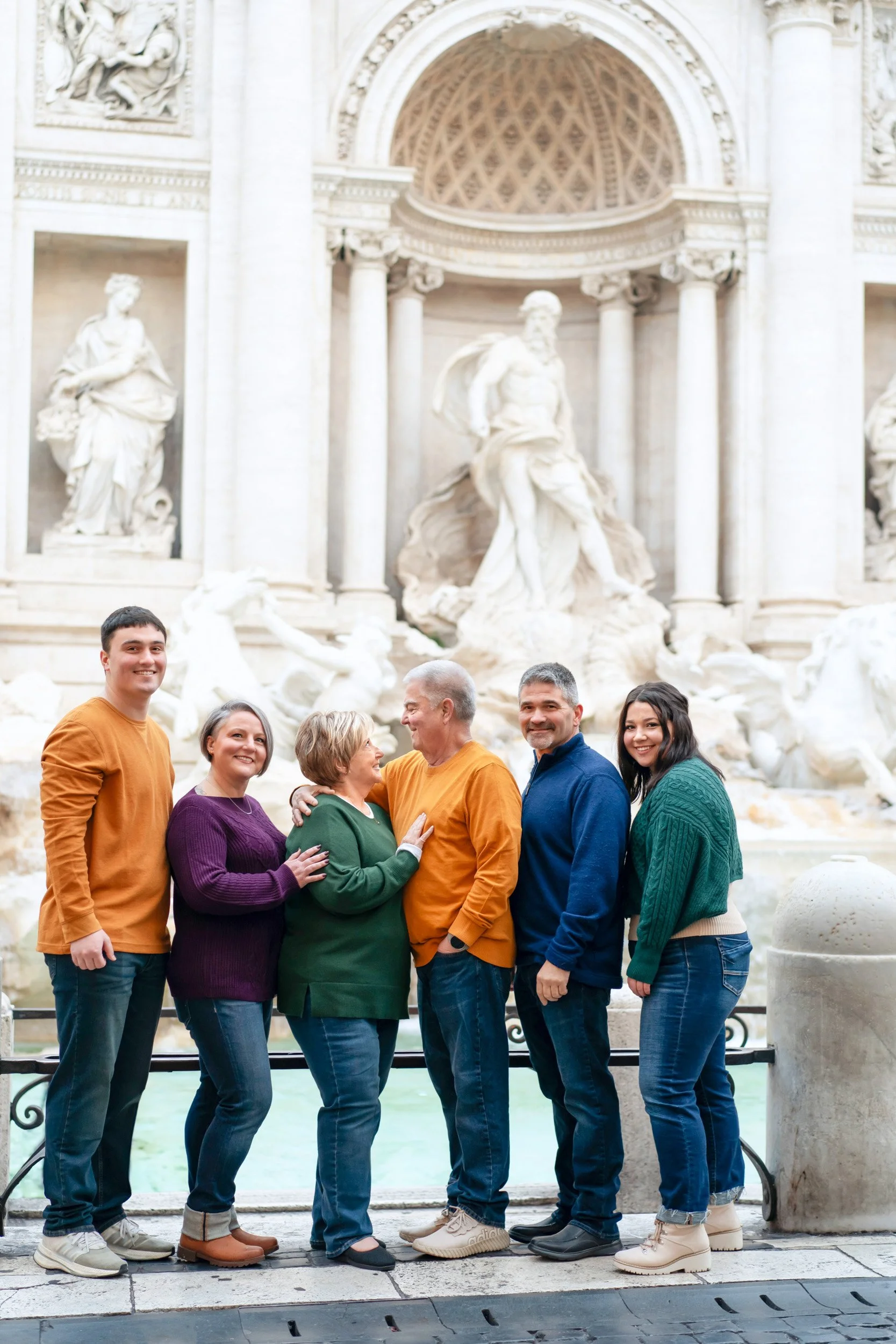 Family of six posing in front of the Trevi Fountain in Rome, Italy. The group includes two young adults, two middle-aged adults, and two older adults, dressed casually in colorful sweaters and jeans, smiling and enjoying the moment.