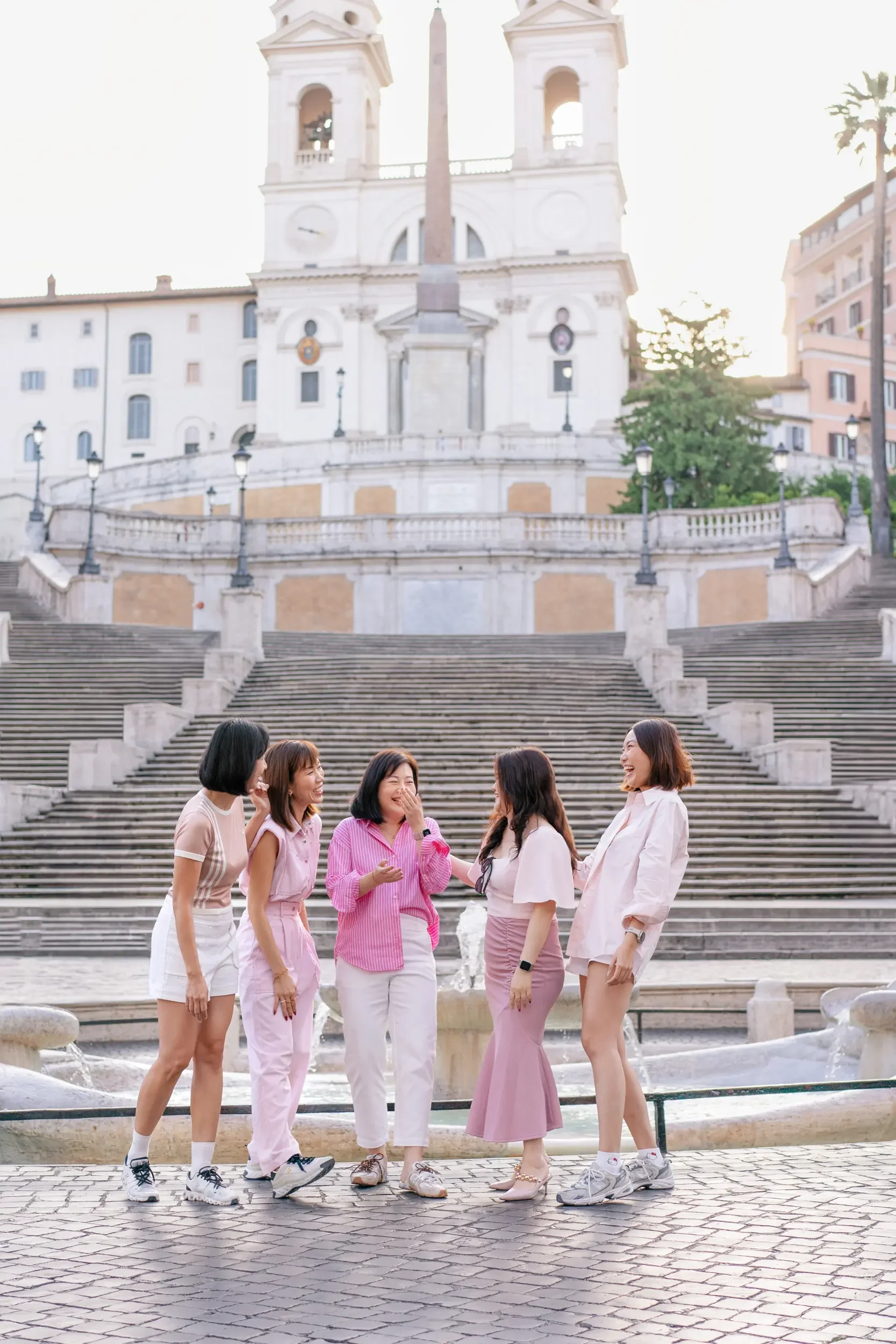 Five women in casual summer outfits laughing and talking in front of a fountain and staircase with a historic building in the background.