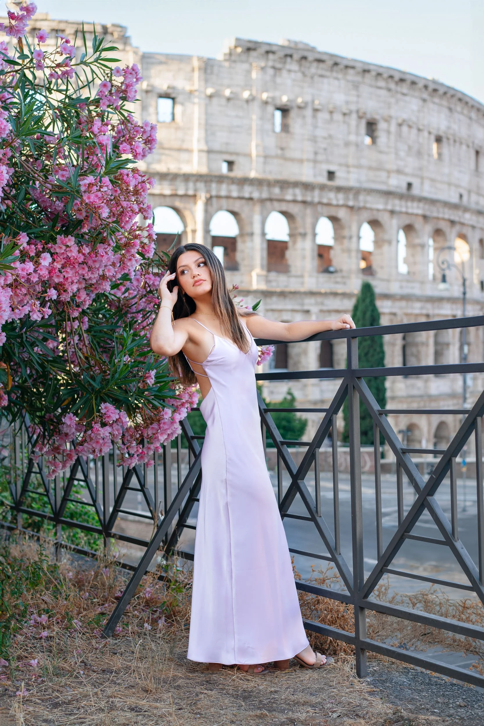 A woman in a long, light purple dress stands beside a pink flowering bush, with the ancient Roman Colosseum in the background.